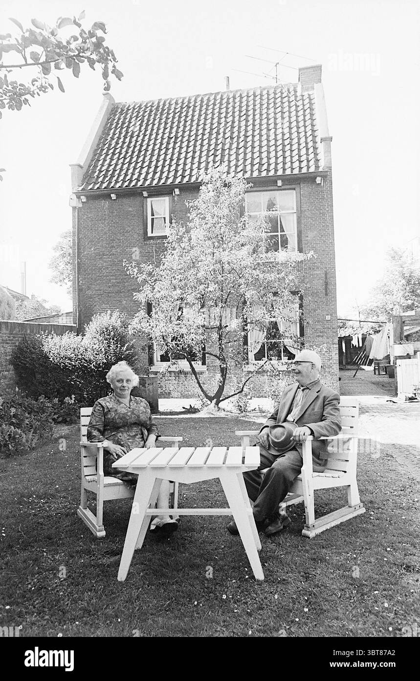 Haus in alten Velsen Häusern und Hausbau, Whizgle News, Dutch Desk, Niederlande, 1950 - 2000 am 29-06-1965. Dies sind die Themen im Bild. In einer ruhigen Umgebung im Freien sitzen zwei ältere Personen bequem an einem weißen Holztisch. Die Frau mit lockigen Haaren und gemustertem Kleid scheint in einem Gespräch mit dem Mann zu sein, der einen Hut und einen hellen Anzug trägt. Beide zeigen ein warmes Lächeln, was ein Gefühl von Gesellschaft und Leichtigkeit andeutet. Hinter ihnen steht ein charmantes Backsteinhaus, das sich durch seine traditionelle Architektur und ein markantes Ziegeldach auszeichnet. Das Fenster des Hauses Stockfoto