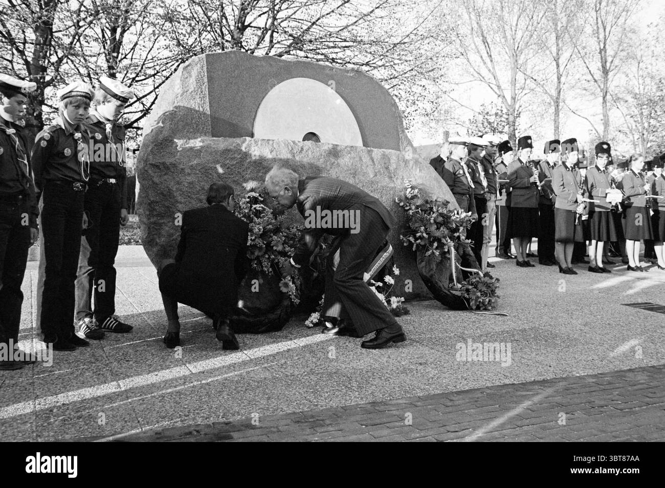 Whizgle News, Dutch Desk, Niederlande, 1950 - 2000 am 04-05-1982. Dies sind die Elemente im Bild. Die Szene fängt einen feierlichen Moment an einer Gedenkstätte ein, die von einem großen, markanten Steindenkmal geprägt ist. Dieses Denkmal verfügt über eine kreisförmige Öffnung, die ihm ein unverwechselbares Aussehen verleiht. Um das Gebäude herum steht eine Gruppe uniformierter Personen in ordentlichen Reihen. Ihre Kleidung, wahrscheinlich militärisch oder zeremoniell, ist von scharfen Kontrasten geprägt, einige tragen Hüte und zeigen verschiedene Abzeichen. Im Vordergrund zwei Individu Stockfoto