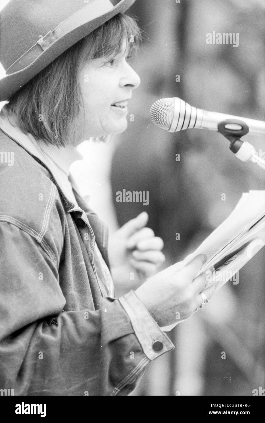 Liberation Puppe H'lem Haarlem the Netherlands, Whizgle News, Dutch Desk, Niederlande, 1950 - 2000 am 04-05-1991. Dies sind die Themen im Bild. In einer lebendigen Umgebung im Freien steht eine Person an einem Mikrofon und nimmt einen animierten Moment ein, während sie spricht oder spielt. Die Person hat schulterlange Haare, die teilweise durch einen stilvollen Hut verdeckt sind, und ihr Ausdruck deutet auf Leidenschaft und Engagement für das Publikum hin. Sie halten einen Stapel Papiere in der einen Hand, die mit der anderen ausdrücken und ein Gefühl der Begeisterung vermitteln. Die Kleidung besteht aus einer Jeansjacke, die lässig und dennoch destilliert wirkt Stockfoto