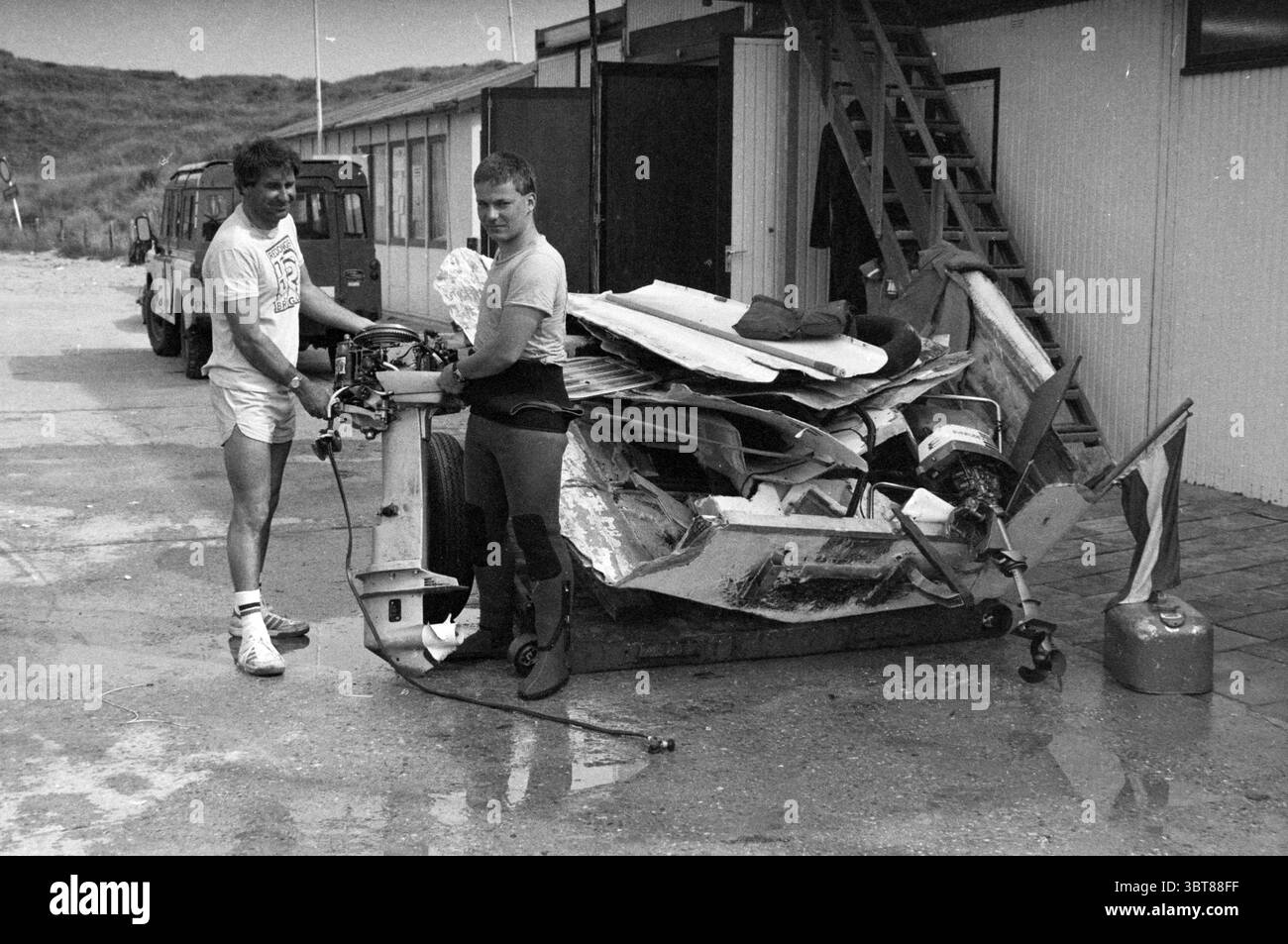Wrack eines Schnellbootes IJmuiden Beach Wrack Entfernung IJmuiden the Netherlands, Whizgle News, Dutch Desk, Niederlande, 1950 - 2000 am 03-07-1983. Diese Themen sind in der Abbildung dargestellt. In einer Szene, die von einer rauen Umgebung im Freien geprägt ist, stehen zwei junge Männer an vorderster Front, die eine Aktivität ausüben, bei der ein beschädigtes oder demontiertes Fahrzeug zu verspüren scheint. Die erste Person, die ein helles T-Shirt und kurze sportliche Shorts trägt, ist leicht links positioniert. Er hat ein entspanntes Auftreten, mit einem leichten Lächeln. Sein Gefährte trägt einen passgenauen Neoprenanzug, was darauf hindeutet, dass er bereit für die WA ist Stockfoto