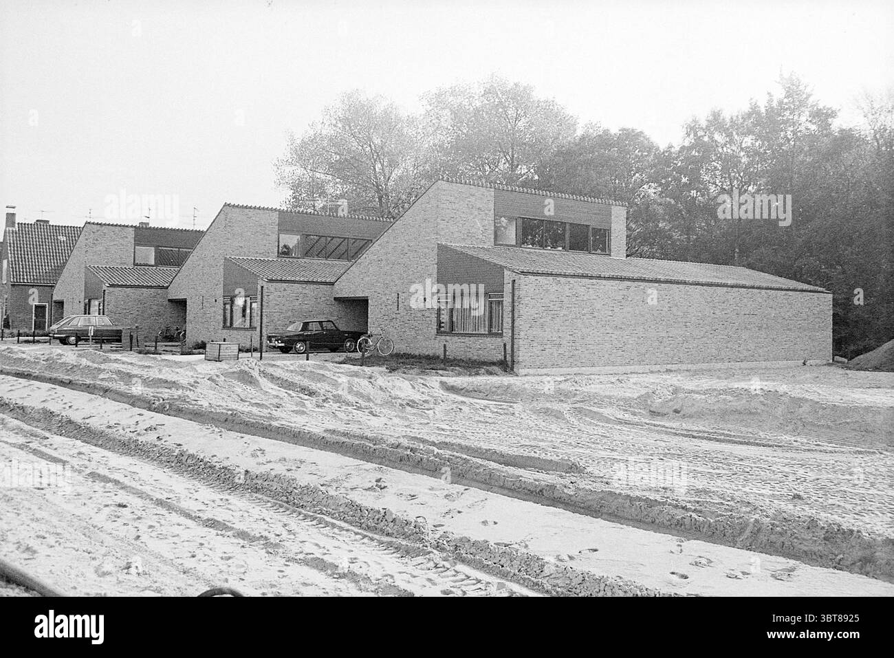 Modell der Häuser Lorentzlaan Heemstede, Whizgle News, Dutch Desk, Niederlande, 1950 - 2000 im Jahr 1972. Diese Themen sind in der Abbildung dargestellt. Die Szene zeigt eine Reihe von modernen, eckigen Häusern, die sich durch ihre flachen Dächer und rechteckigen Formen auszeichnen. Die Gebäude sind aus hellen Ziegelsteinen gebaut, was ihnen ein sauberes und minimalistisches Aussehen verleiht. Große Fenster ermöglichen einen Blick auf die Innenräume und lassen natürliches Licht in Kontrast zu den gedämpften Außentönen. Im Vordergrund erstreckt sich ein sandiger Bereich über die Szene, der auf Bau- oder Landschaftsarbeiten hinweist. Reifenspuren weben Stockfoto