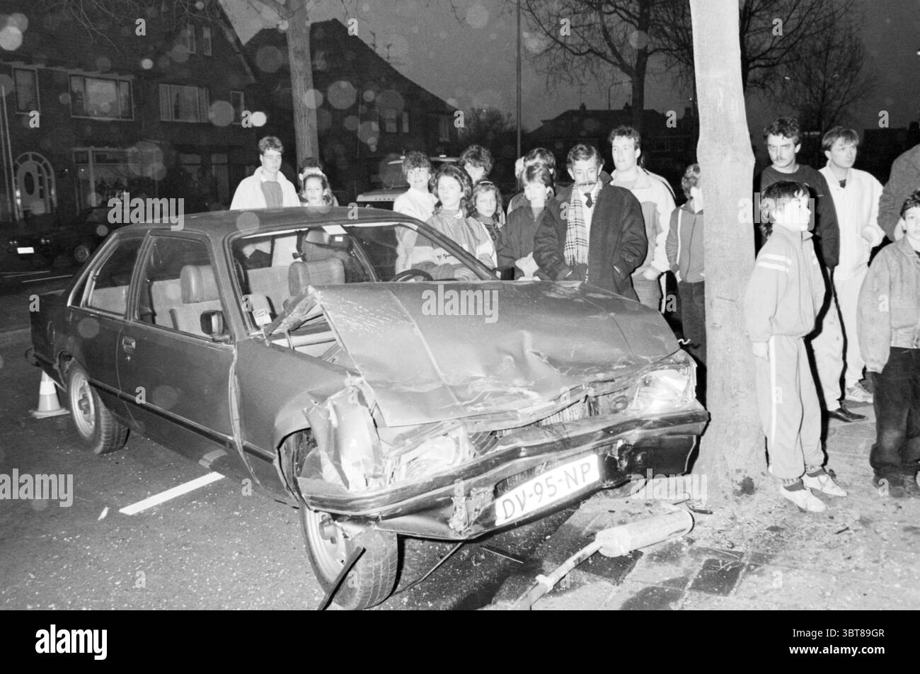Autounfall Feuerwehr bei einem Autounfall auf der Straße., Whizgle News, Dutch Desk, Niederlande, 1950 - 2000 am 03-1986. Die Abbildung zeigt diese Themen. In einer schwach beleuchteten Stadt versammelt sich eine Gruppe von Zuschauern um ein beschädigtes Auto, das in einen Baum gestürzt ist. Das Fahrzeug, eine alte Limousine, weist erhebliche Schäden an der Front auf, die durch zerknittertes Metall und zersplittertes Glas verursacht wurden. Die Szene zeichnet sich durch die gedämpften nächtlichen Töne aus, wobei dunkle Schattierungen die Visualisierung dominieren, unterbrochen durch das gelegentliche reflektierende Leuchten der nahen Straßenlaternen. Die Gruppe der Menschen, vorwiegend Kinder und Jugendliche, steht in der Nähe Stockfoto