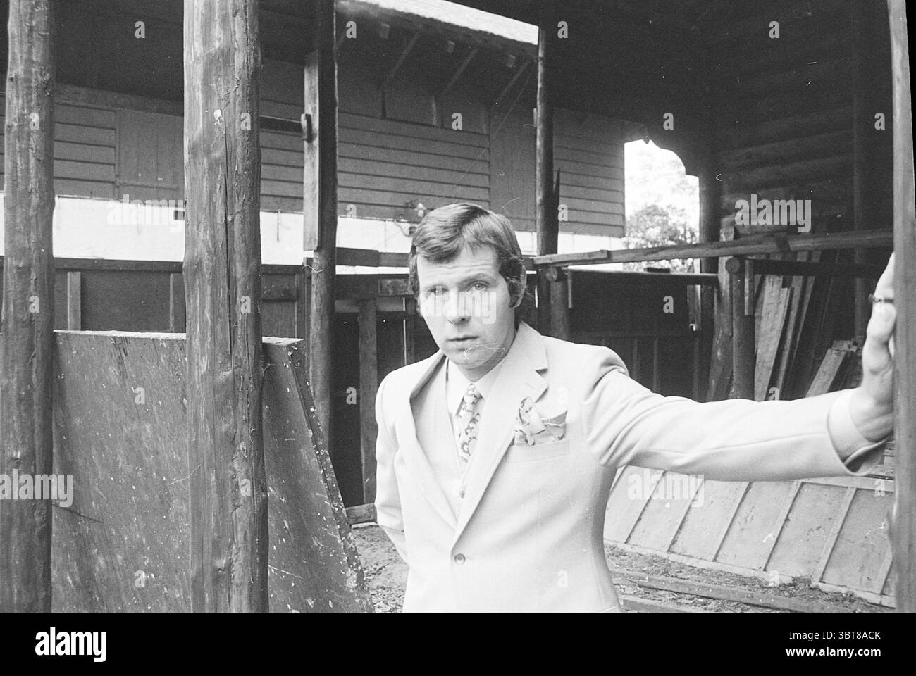 Fons Post vor der Scheune eines Schweizer Bauernhofs Personen, Whizgle News, Dutch Desk, Niederlande, 1950 - 2000 am 26-05-1971. Die Abbildung zeigt diese Themen. In der Szene steht ein Mann selbstbewusst in einer rustikalen Umgebung, die von verwitterten Holzkonstruktionen geprägt ist, die an ein verlassenes oder baufälliges Gebäude erinnern. Er trägt einen hellen Anzug, der auffällig mit dem dunklen, alten Holz um ihn herum kontrastiert. Der Anzug verfügt über ein scharfes Revers und eine dekorative Blume auf der linken Seite, die seinem ansonsten ernsten Ausdruck einen Hauch von Eleganz verleiht. Sein Haar ist sauber gekämmt und sein Blick Stockfoto