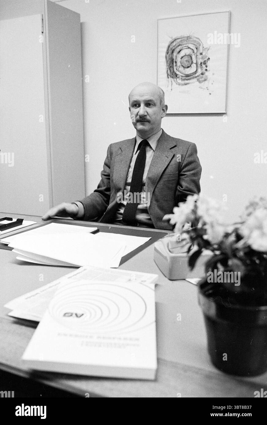 Porträt eines Gentleman hinter dem Schreibtisch mit Buch über Energiesparen, Whizgle News, Dutch Desk, Niederlande, 1950 - 2000 auf 01-1979. Das Bild enthält diese Themen. In einer hellen Büroumgebung sitzt ein Mann selbstbewusst am Schreibtisch. Sein Aussehen ist formell, mit einem maßgeschneiderten grauen Anzug mit einem knusprigen weißen Hemd und einer dunklen Krawatte, die Professionalität vermittelt. Sein Ausdruck ist ruhig und komponiert, was auf ein nachdenkliches Verhalten hindeutet. Der Schreibtisch ist ordentlich organisiert, mit ein paar Blättern Papier und einem Blumentopf, der mit kleinen, zarten Blumen geschmückt ist. Die Blüten sorgen für einen Hauch Wärme und schaffen Stockfoto
