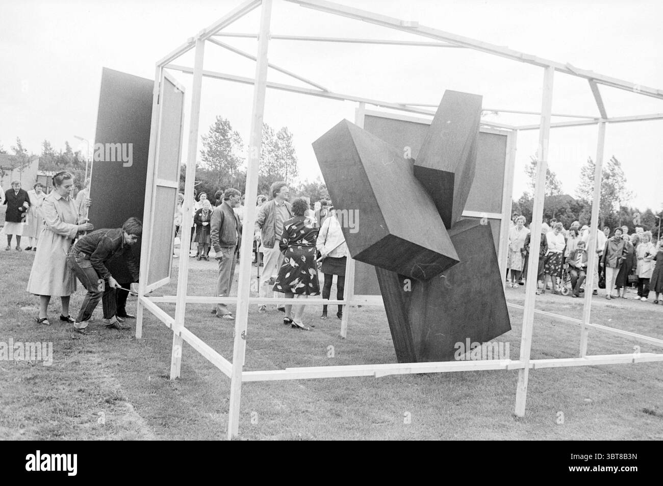 Enthüllung der Statue von Sancta Maria Noordwijkerhout Revelations Noordwijkerhout, Whizgle News, Dutch Desk, Niederlande, 1950 - 2000 am 26-06-1984. Diese Themen sind in der Abbildung dargestellt. Im Freien versammelt sich eine Gruppe von Menschen um eine auffällige Skulptur aus großen geometrischen Formen. Die Skulptur scheint aus dunklen, festen Materialien zu bestehen, mit drei bedeutenden kubischen Formen, die in verschiedenen Winkeln positioniert sind, was ihr ein Gefühl von Dynamik und Gewicht verleiht. Die Struktur ist von einem Skelettrahmen aus weißem Metall umschlossen, der einen starken Kontrast zum dunkleren bildet Stockfoto