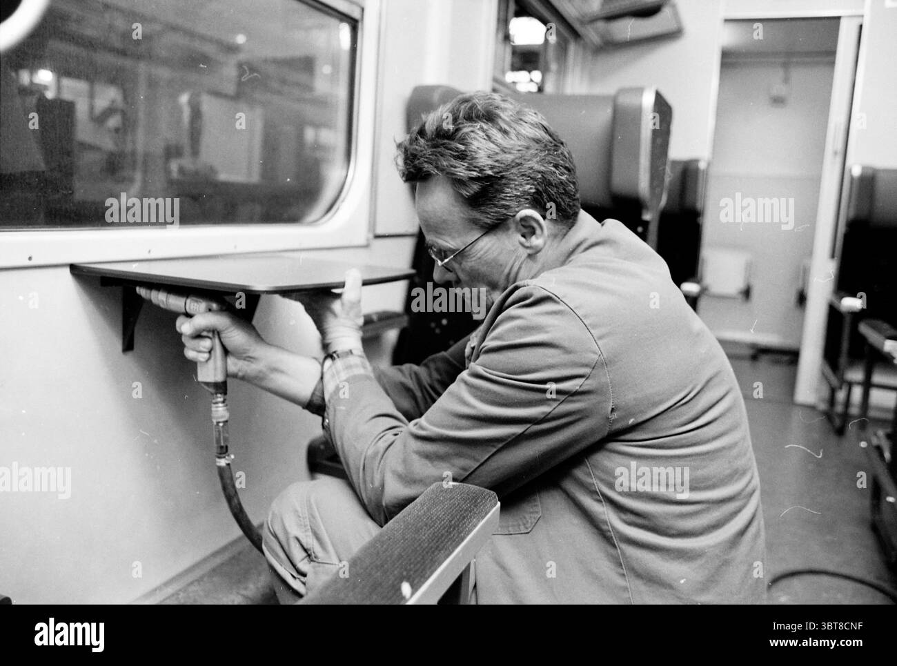 Collision Department Ned Train, Whizgle News, Dutch Desk, Niederlande, 1950 - 2000 am 10-04-2002. Dies sind die Themen im Bild. Ein Mann sitzt auf einer Bank in einem Zugabteil, in seiner Arbeit vertieft. Er neigt sich leicht nach vorne und konzentriert sich intensiv auf eine Aufgabe, bei der ein Werkzeug in seinen Händen liegt. Die Umgebung ist schwach beleuchtet, was eine etwas düstere Atmosphäre schafft, mit Schatten, die entlang der Wände und der Oberfläche des Abteils spielen. Er trägt ein helles, zweckmäßiges Outfit, das den praktischen Look abrundet. Das Innere des Fachs ist eine Mischung aus gedämpften Farben – Braun und Grau Stockfoto