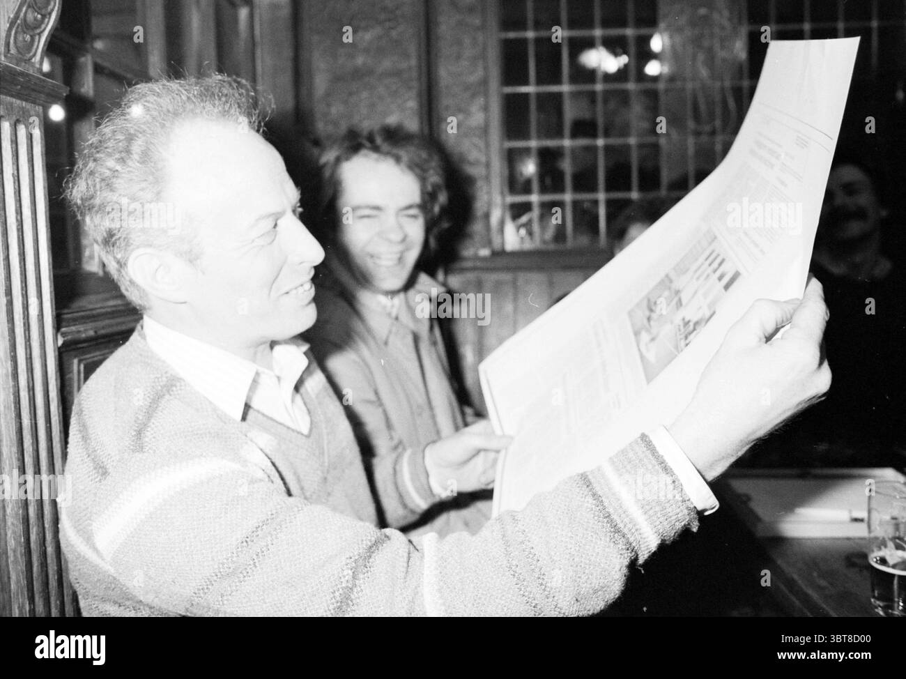 Cor Boos 10 Jahre bei HD Haarlems Dagblad etc. Und IJmuider Courant and Koeri, Whizgle News, Dutch Desk, Niederlande, 1950 - 2000 am 02-02-1986. Dies sind die Elemente im Bild. In einem gemütlichen Innenbereich sind zwei Personen in einem unbeschwerten Moment beschäftigt. Eine Person, die links sitzt, hat einen fröhlichen Ausdruck, als sie ein großes Blatt Papier untersucht, das er in beiden Händen hält. Er hat graue Haare und trägt einen hellen, gestreiften Pullover. Seine Eigenschaften deuten auf Unterhaltung oder Neugier hin, da er sich intensiv auf die Zeitung konzentriert. Zu seiner Rechten wird ein anderer Mann mit einem breiten Lächeln gesehen. Stockfoto