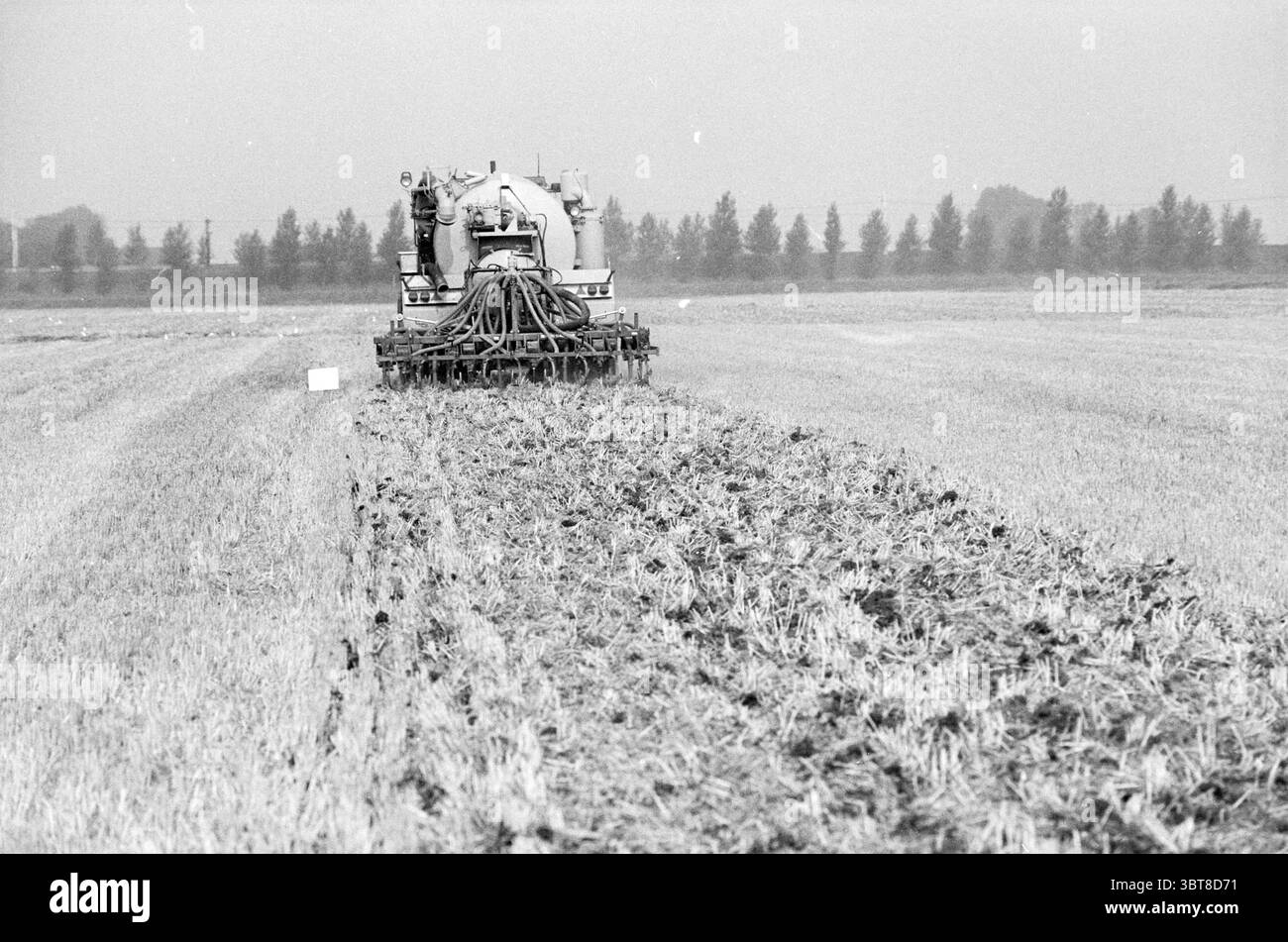 Demonstration des Stoppelinjektors Nieuw Vennep Nieuw-Vennep, Whizgle News, Dutch Desk, Niederlande, 1950 - 2000 am 27-08-1990. Diese Themen sind in der Abbildung dargestellt. Die Szene fängt eine riesige landwirtschaftliche Landschaft ein, die durch Flecken von sanften Feldern gekennzeichnet ist, die sich bis in den Horizont erstrecken. Ein großes, mechanisiertes Landwirtschaftsfahrzeug dominiert den Vordergrund und ist für spezifische Aufgaben in der Erntegutwirtschaft konzipiert. Sein Gehäuse hat ein robustes, industrielles Erscheinungsbild und verfügt über verschiedene Rohre und Anbaugeräte, die nach außen ragen, was auf seine Rolle in der landwirtschaftlichen Verarbeitung hinweist. Die Farbpalette ist überwiegend stummgeschaltet, W Stockfoto