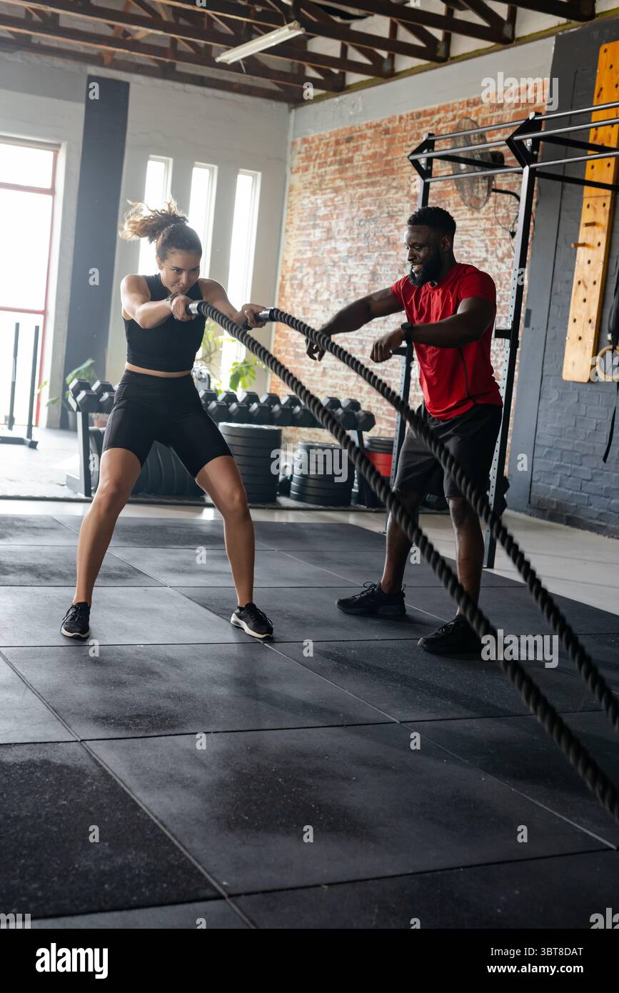 Verschiedene Trainer und Kunden verwenden Kampfseile, während sie im Fitnessstudio in der Nähe des Hantelständers die Form leiten Stockfoto