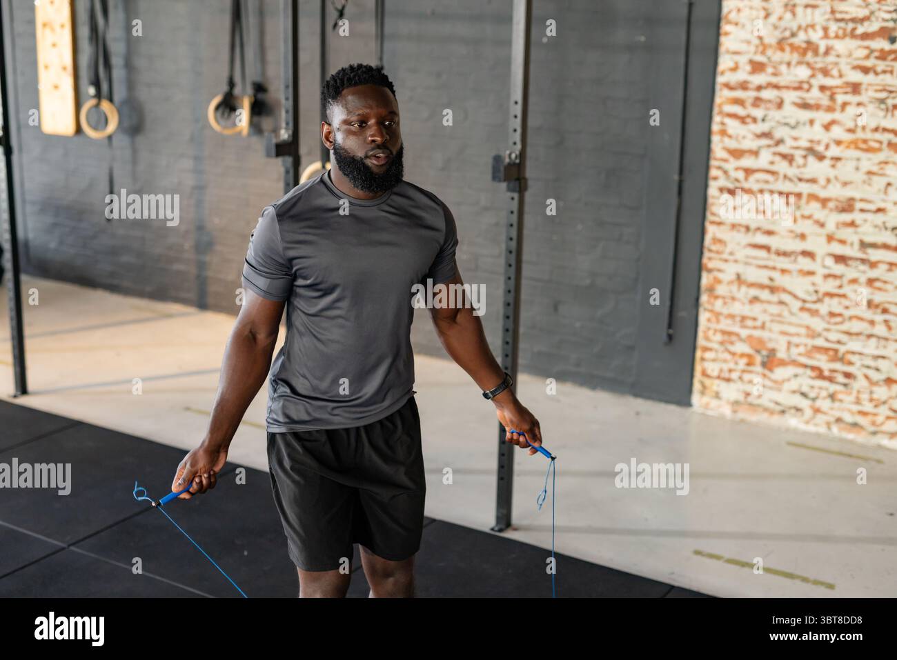 Afroamerikanischer Mann, der ein Seil mit blauen Griffen auf Matten im Fitnessstudio mit Turnringen überspringt Stockfoto