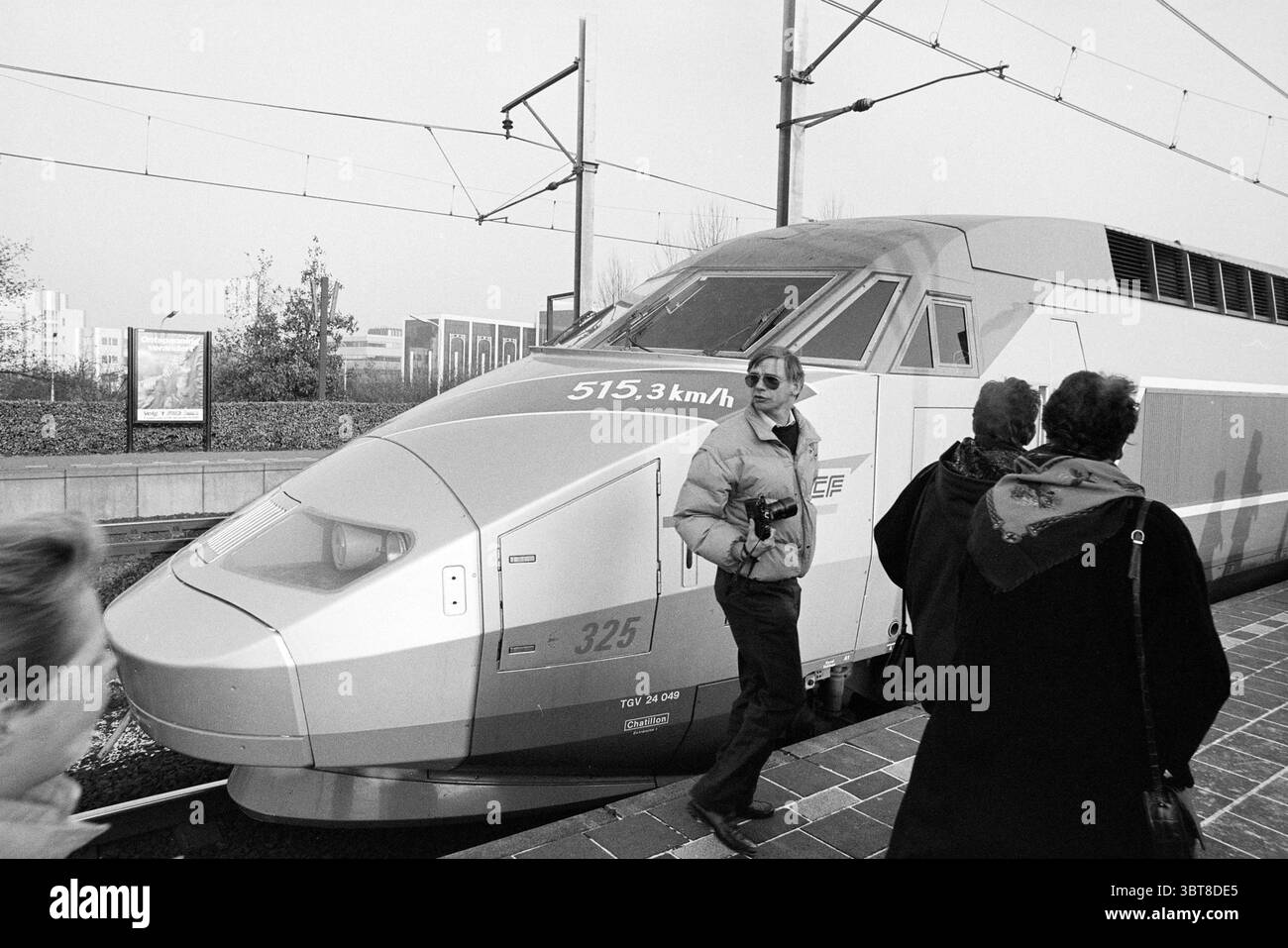 Ankunft TGV Hoofddorp the Netherlands, Whizgle News, Dutch Desk, Niederlande, 1950 - 2000 am 24-11-1993. Das Bild enthält diese Themen. In einem belebten Bahnhof steht ein eleganter, moderner Hochgeschwindigkeitszug auf dem Bahnsteig, dessen aerodynamische Form das modernste Design widerspiegelt. Die Oberfläche des Zuges besteht aus einer Mischung aus weiß und Metallic-Grau, die mit leuchtenden roten Streifen entlang der Länge betont werden, was auf Geschwindigkeit und Effizienz hinweist. Die Zahlen auf der Seite zeigen eine beeindruckende Geschwindigkeit an, die die fortschrittlichen Fähigkeiten des Zuges unterstreicht. Die Atmosphäre ist lebhaft, mit einer kleinen Gruppe Stockfoto
