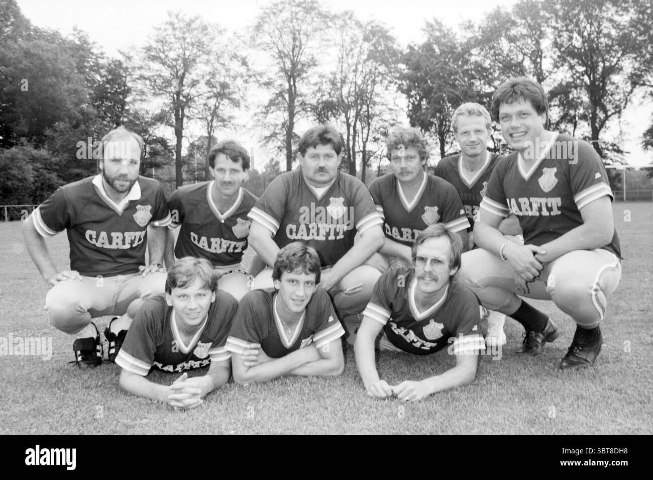Teamfoto CarFit Teams, Whizgle News, Dutch Desk, Niederlande, 1950 - 2000 am 27-08-1987. Dies sind die Elemente im Bild. Eine Gruppe von zehn Individuen versammelt sich in einer grasbewachsenen Outdoor-Umgebung, wahrscheinlich auf einem Sportplatz. Sie tragen passende Trikots, die auf der linken Seite ein Wappen oder Emblem tragen. Die Trikots haben eine tiefe Farbe, möglicherweise kastanienbraun oder dunkelrot, mit kontrastierenden weißen oder helleren Akzenten. Die Gruppe ist in zwei Reihen angeordnet: In der hinteren Reihe stehen fünf Männer, während die erste Reihe aus vier Männern besteht, die kniend oder sitzend sind, wobei einer im Cent liegt Stockfoto