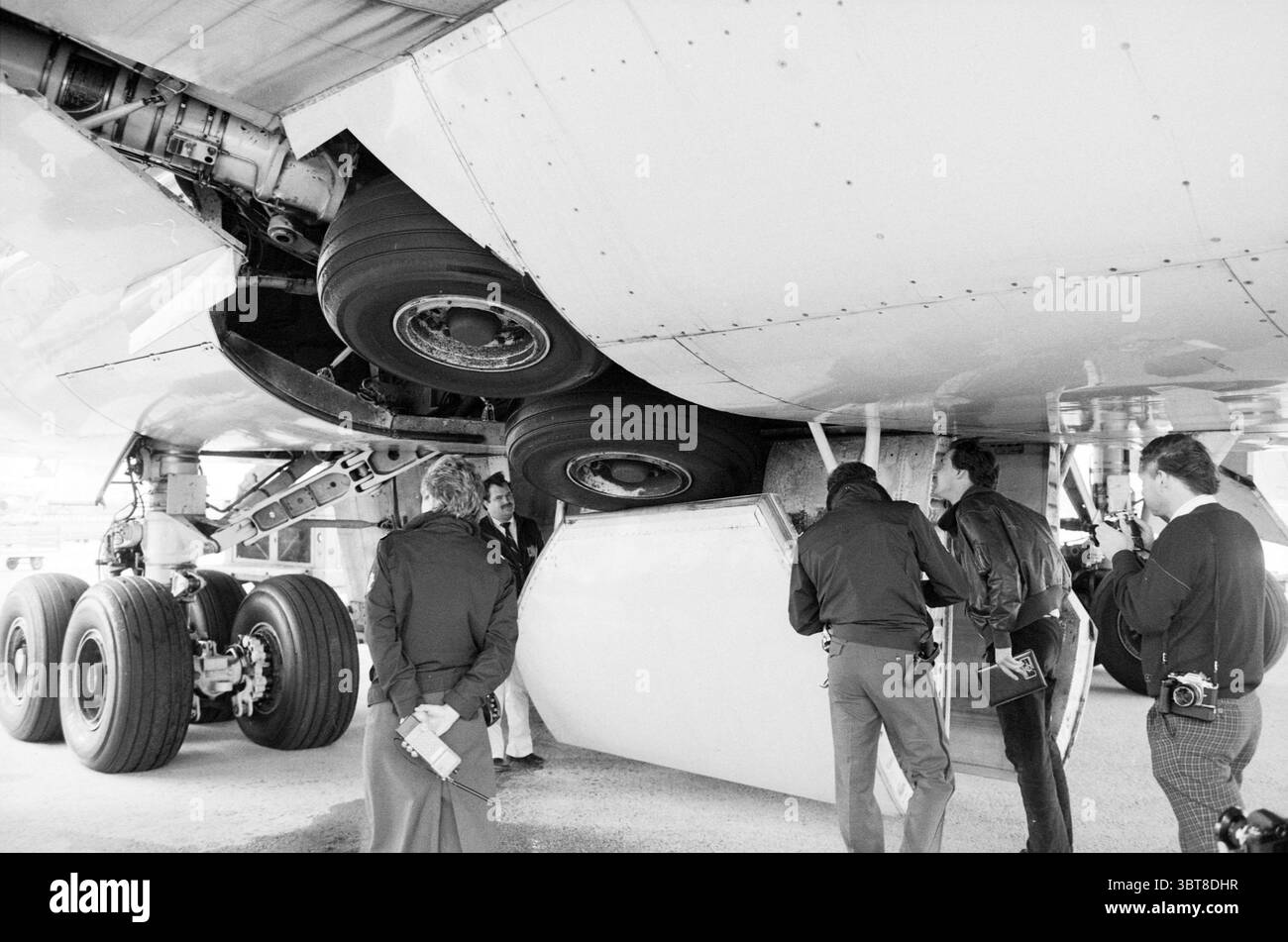 Blockierte Räder anthony fokker Airplanes, Whizgle News, Dutch Desk, Niederlande, 1950 - 2000 am 03-05-1988. Diese Themen sind in der Abbildung dargestellt. Die Szene zeigt eine Gruppe von Individuen, die sich unter dem Bauch eines großen Flugzeugs versammelt haben. Das Flugzeug ist überwiegend weiß, mit Anklängen von Metallic-Grau, die dezentes Licht reflektieren. Unterhalb des Flugzeugs sind mehrere riesige Reifen zu sehen, zusammen mit Teilen des Fahrwerks, was der Szene ein Gefühl von Skala und Kraft verleiht. Rund um das Flugzeug ist eine Mischung von Menschen mit Beobachtung und Diskussion beschäftigt. Einige scheinen in dunklerem, formelleren Akt gekleidet zu sein Stockfoto