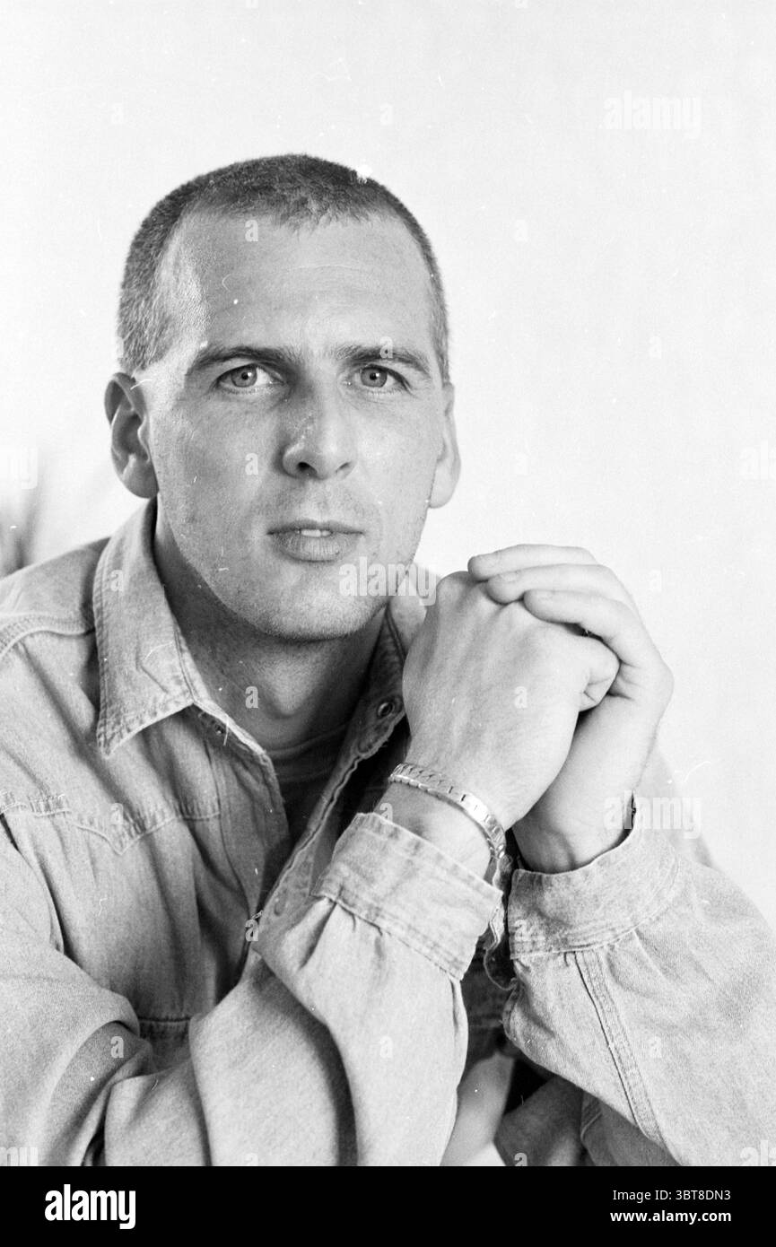 Fred Kuipers Velserbroek, Whizgle News, Dutch Desk, Niederlande, 1950 - 2000 am 13-05-1991. Diese Themen sind in der Abbildung dargestellt. In dieser Szene wird ein Mann in einer entspannten, aber aufmerksamen Pose positioniert. Er sitzt mit aneinander gefalteten Händen, was auf ein Gefühl der Kontemplation oder Konzentration hindeutet. Sein Kopf ist leicht geneigt, und er blickt direkt auf den Zuschauer mit durchdringenden blauen Augen, die eine nachdenkliche und ernste Stimmung vermitteln. Der Mann hat eine eng geschnittene Frisur, die ihm ein klares und raffiniertes Aussehen verleiht. Er trägt eine helle Denim-Jacke, die noch lässiger ist Stockfoto