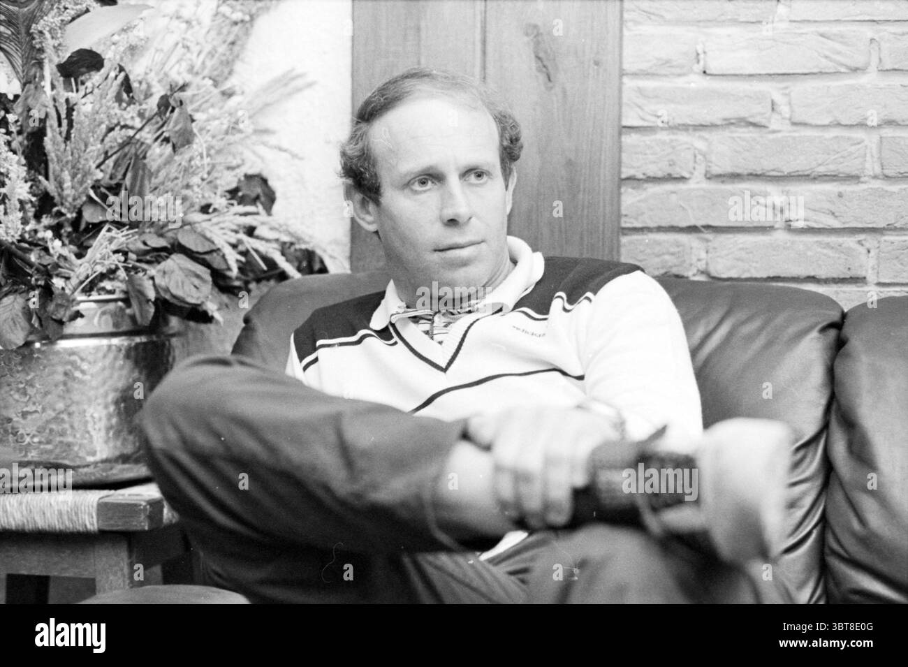 Portrait Eddy Treitel AZ 67 Alkmaar Portraits Football Alkmaar, Whizgle News, Dutch Desk, Niederlande, 1950 - 2000 am 01-05-1981. Das Bild enthält diese Themen. Ein Mann sitzt bequem auf einer dunklen Ledercouch und strahlt ein entspanntes, aber besinnliches Verhalten aus. Seine Kleidung trägt einen weißen Pullover mit schwarzen und marineblauen Streifen und eine dunkle Hose. Er legt einen Arm auf die Rückseite der Couch und hält sein anderes Bein über dem Knie, was auf eine lässige Haltung hindeutet. Das Zimmer hat ein gemütliches Ambiente mit warmen Tönen, die durch die Holzelemente im Hintergrund erzeugt werden, einschließlich einer Holzplatte im Hintergrund Stockfoto