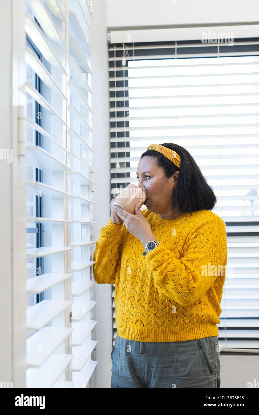 Afroamerikanische Erwachsene Frau, die an den Fensterläden steht und einen Becher in einem Senfpullover hält, Kopierraum Stockfoto