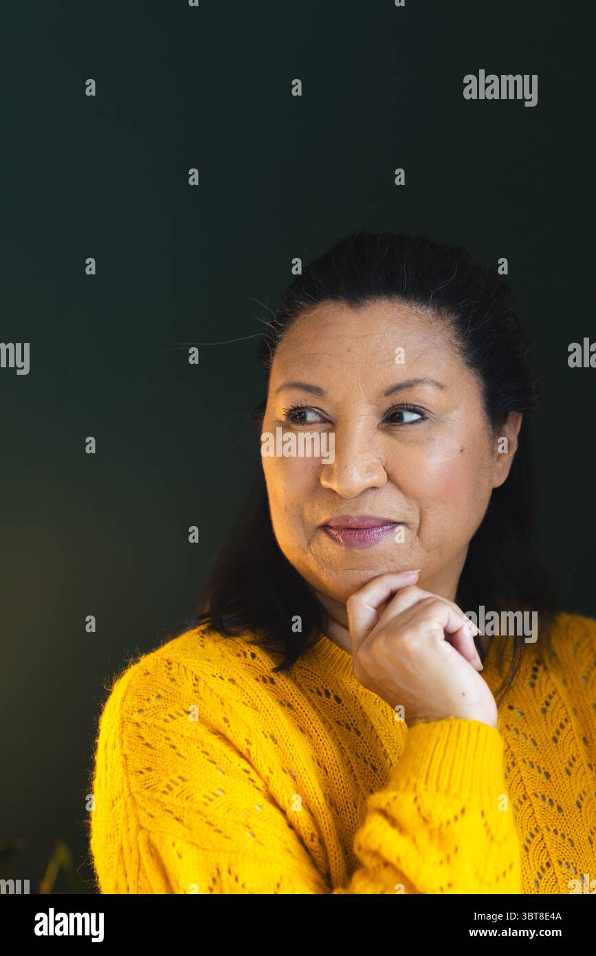 Frau mittleren Alters, die das Kinn auf der Hand an der dunkelgrünen Wand anliegend, mit gelbem Pullover, Kopierraum Stockfoto