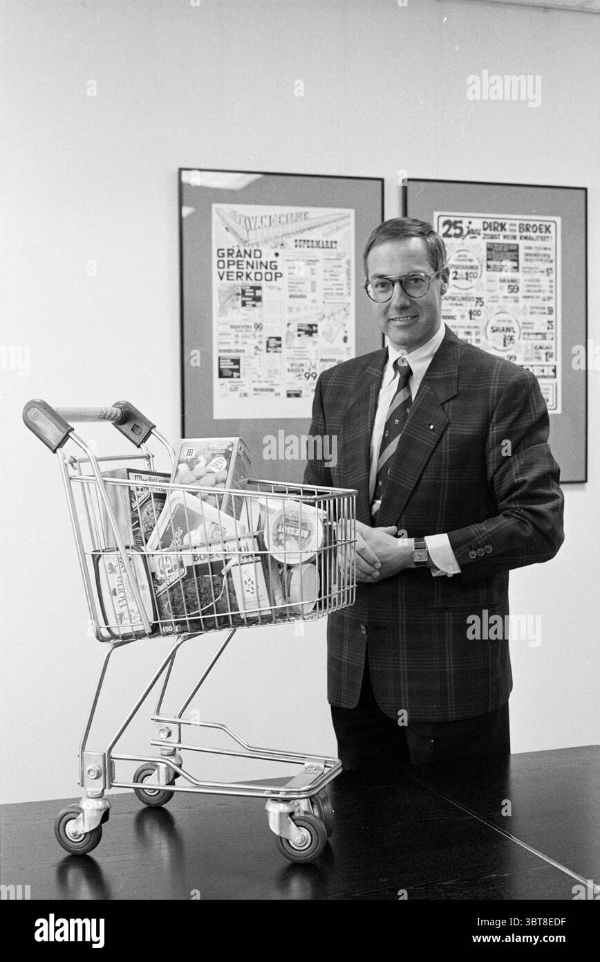 Dirk v.d. Broek Hauptsitz Beukenhorst Supermarket, Whizgle News, Dutch Desk, Niederlande, 1950 - 2000 am 16.03.1990. Das Bild enthält diese Themen. Die Szene zeigt einen Mann mittleren Alters, der neben einem Einkaufswagen mit Lebensmitteln steht. Er trägt eine karierte Anzugsjacke, ein Hemd mit Kragen und eine Krawatte, die ein formelles, aber ansprechendes Aussehen vermittelt. Der Mann hat kurze, ordentlich gekämmte Haare und trägt eine Brille, was sein professionelles Verhalten verstärkt. Der Warenkorb, metallisch und elegant, steht im Kontrast zu den Artikeln im Inneren, die aus verschiedenen verpackten Waren bestehen, was auf eine Tippfunktion hindeutet Stockfoto