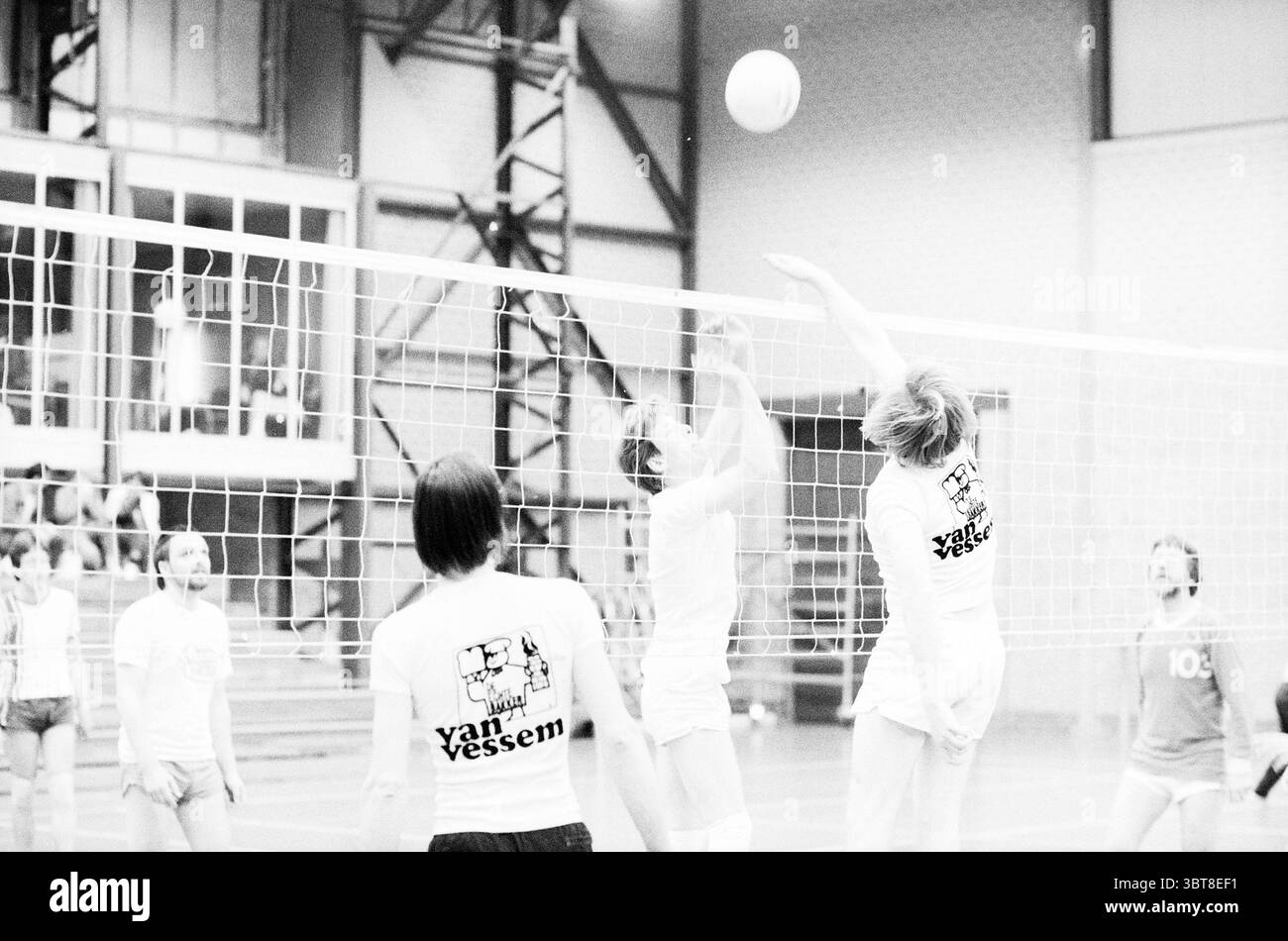 Volley-Ball, Whizgle News, Dutch Desk, Niederlande, 1950 - 2000 am 04-1985. Das Bild enthält diese Themen. Die Szene fängt ein Indoor-Volleyballspiel ein, das sich durch eine dynamische und energiegeladene Atmosphäre auszeichnet. Der Hof ist von hohen Decken und großen Fenstern umgeben, die weiches, diffuses Tageslicht hereinlassen und den Raum mit einem hellen, aber sanften Licht beleuchten. Im Vordergrund stehen zwei Spieler in einem Moment der Action. Der eine ist Mittelsprung, der hoch ansteigt, um gegen einen eintretenden Volleyball zu blockieren, dessen Fokus in der Körpersprache deutlich wird. Dieser Spieler trägt ein weißes Trikot, während er Stockfoto