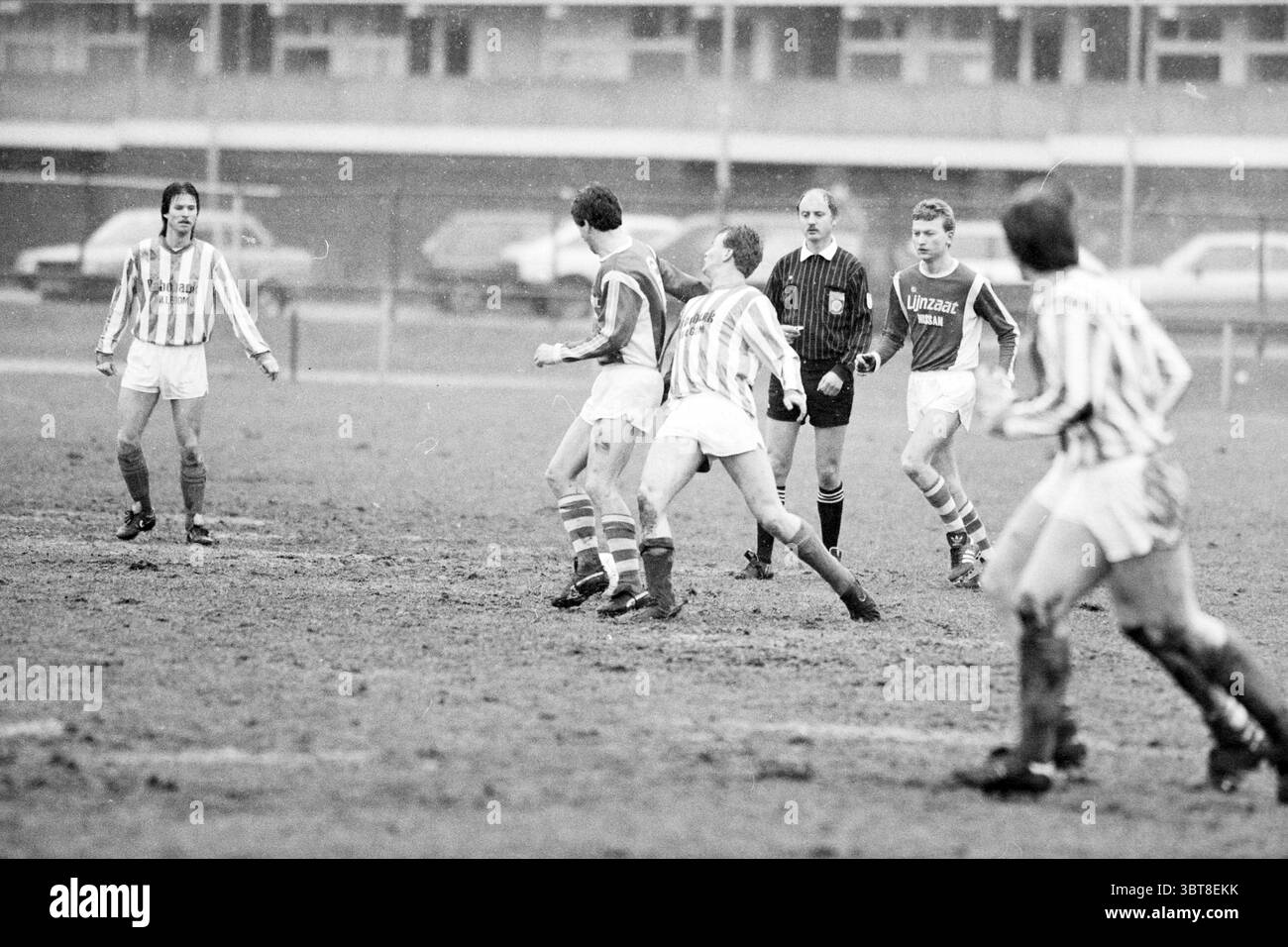 DIO - Hillegom Football DIO, Whizgle News, Dutch Desk, Niederlande, 1950 - 2000 am 13-03-1988. Dies sind die Elemente im Bild. Die Szene fängt die Intensität eines Fußballspiels in einer etwas düsteren, bewölkten Atmosphäre ein. Eine Gruppe von Spielern, die in abwechselnd gestreiften Trikots gekleidet sind – eine Mannschaft in horizontalen rot-weißen Streifen und die andere in vertikalem Blau –, spielt auf einem matschigen Spielfeld aktiv. Der Boden ist sichtbar abgenutzt, mit kaum sichtbaren Rasenflächen, was auf schwierige Wetterbedingungen hindeutet, die das Spiel beeinflusst haben. Im Vordergrund stehen zwei Spieler aus Rot und weiß. Stockfoto