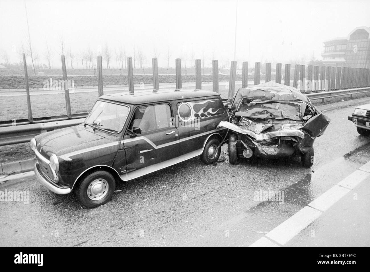 Autounfall aufgrund rutschiger Bedingungen, Whizgle News, Dutch Desk, Niederlande, 1950 - 2000 am 02-1987. Diese Themen sind in der Abbildung dargestellt. Die Szene zeigt einen grauen und bewölkten Tag, an dem ein kleines Oldtimerfahrzeug, das sich durch sein kompaktes Design und einzigartige Flammaufkleber an der Seite auszeichnet, links geparkt wird. Die Farbe des Fahrzeugs ist gedämpft und kontrastiert leicht mit den dunkleren Farben in der Umgebung. Neben dem Oldtimer befindet sich ein deutlich größeres, stark beschädigtes Fahrzeug. Das hintere Ende ist zerfetzt und zerknittert, was auf eine schwere Kollision hindeutet. Die metallische Oberfläche Stockfoto