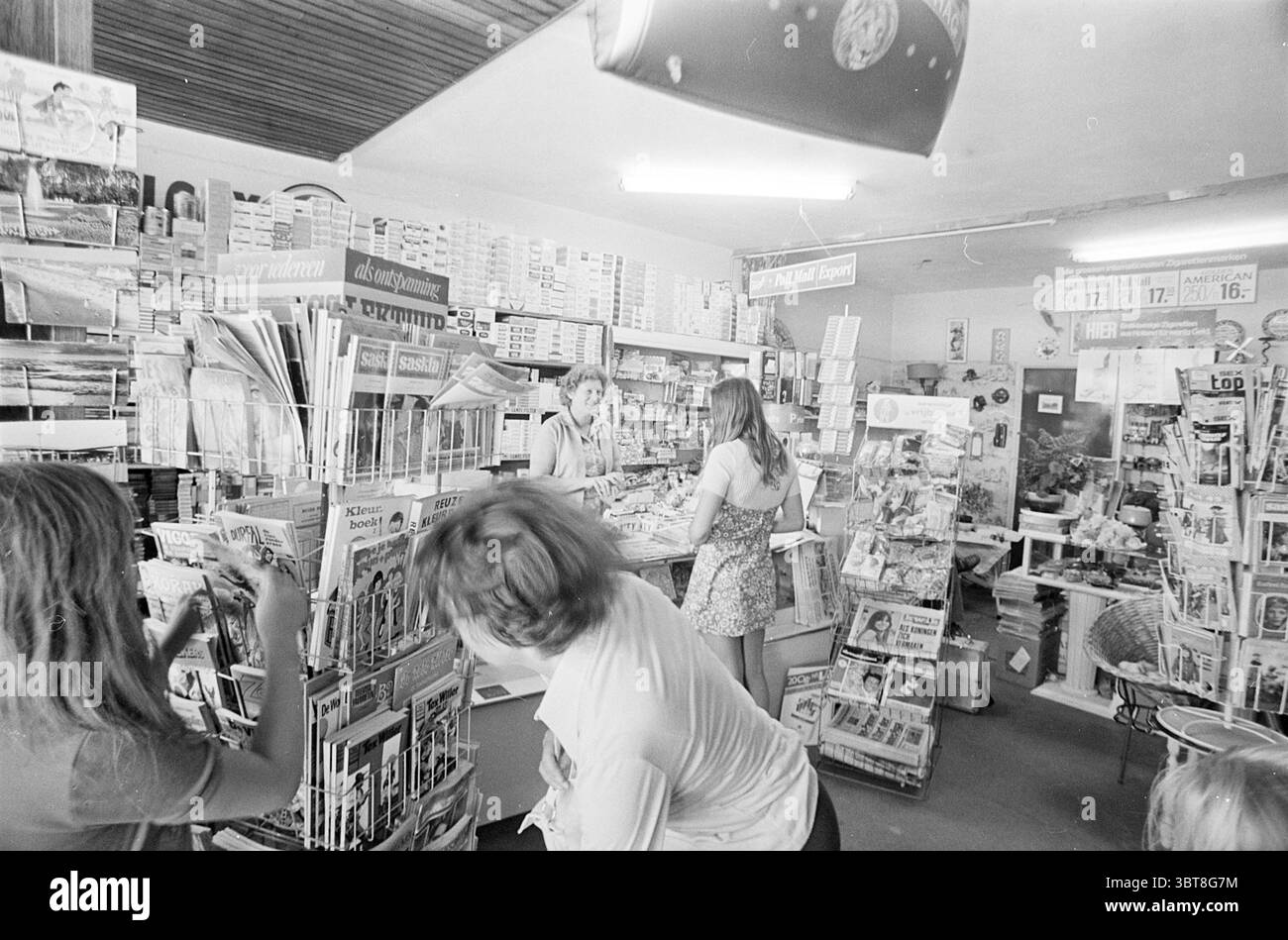 Magazin und Tabakladen, Whizgle News, Dutch Desk, Niederlande, 1950 - 2000 im Jahr 1974. Das Bild enthält diese Themen. Die Szene spielt in einem kleinen, geschäftigen Geschäft, das mit verschiedenen Drucksachen gefüllt ist. Der Innenraum hat eine etwas nostalgische Atmosphäre, die durch den Schwarz-weiß-Ton verstärkt wird. Die Atmosphäre ist lebhaft, mit mehreren Personen, die an verschiedenen Aktivitäten teilnehmen. Links sieht eine Frau aus, als ob sie durch ein Regal voller Zeitschriften stöbert, ihr Haar leicht zerzaust und ein lässiges Outfit trägt. Das Gestell ist farbenfroh und mit lebhaften Magazinumschlägen gefüllt, die sich in einem Kontrast zur monochromen Umgebung befinden. Stockfoto