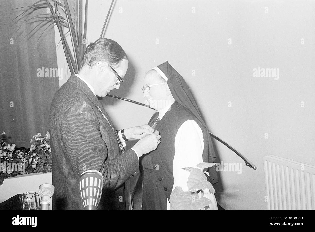 Hommage an Schwester Helena. Homage, Whizgle News, Dutch Desk, Niederlande, 1950 - 2000 am 24-10-1968. Das Bild enthält diese Themen. Die Szene fängt einen feierlichen und intimen Moment zwischen zwei Personen ein, wahrscheinlich in einem formellen Umfeld. Eine Person, die in einem maßgeschneiderten Anzug mit einem knusprigen Hemd und einer Krawatte gekleidet ist, befestigt sanft eine Medaille oder ein Abzeichen auf der Kleidung einer anderen Person. Diese zweite Person trägt eine traditionelle Nonnengewohnheit, die durch ein langes Kleid und einen unverwechselbaren Kopfschmuck gekennzeichnet ist, der ein Gefühl von Hingabe und Formalität vermittelt. Das Ambiente wirkt dezent, aber elegant, mit einem Stockfoto