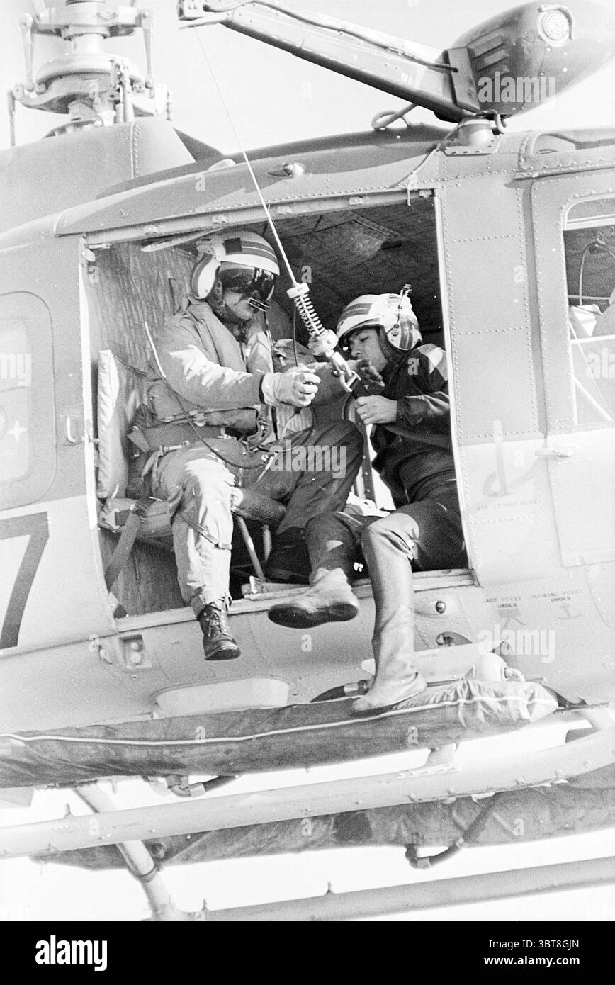 Rettungsübung von Piloten aus der Nordsee mit SAR Helikoptern Air Force and Alarm, Whizgle News, Dutch Desk, Niederlande, 1950 - 2000 am 02-10-1975. Diese Themen werden in der Abbildung angezeigt. Die Szene fängt einen dynamischen Moment an Bord eines Militärhubschraubers ein. Zwei Personen sind besonders hervorgehoben, beide tragen Schutzausrüstung einschließlich Helmen und Kopfhörern, was auf eine Umgebung hindeutet, die auf dem Spiel steht. Eine Person sitzt im Helikopter und konzentriert sich auf eine Aufgabe, während die andere auf halbem Weg aus dem Helikopter positioniert ist, scheinbar bereit, einzugreifen oder zu helfen. Das Innere ist voll von mir Stockfoto