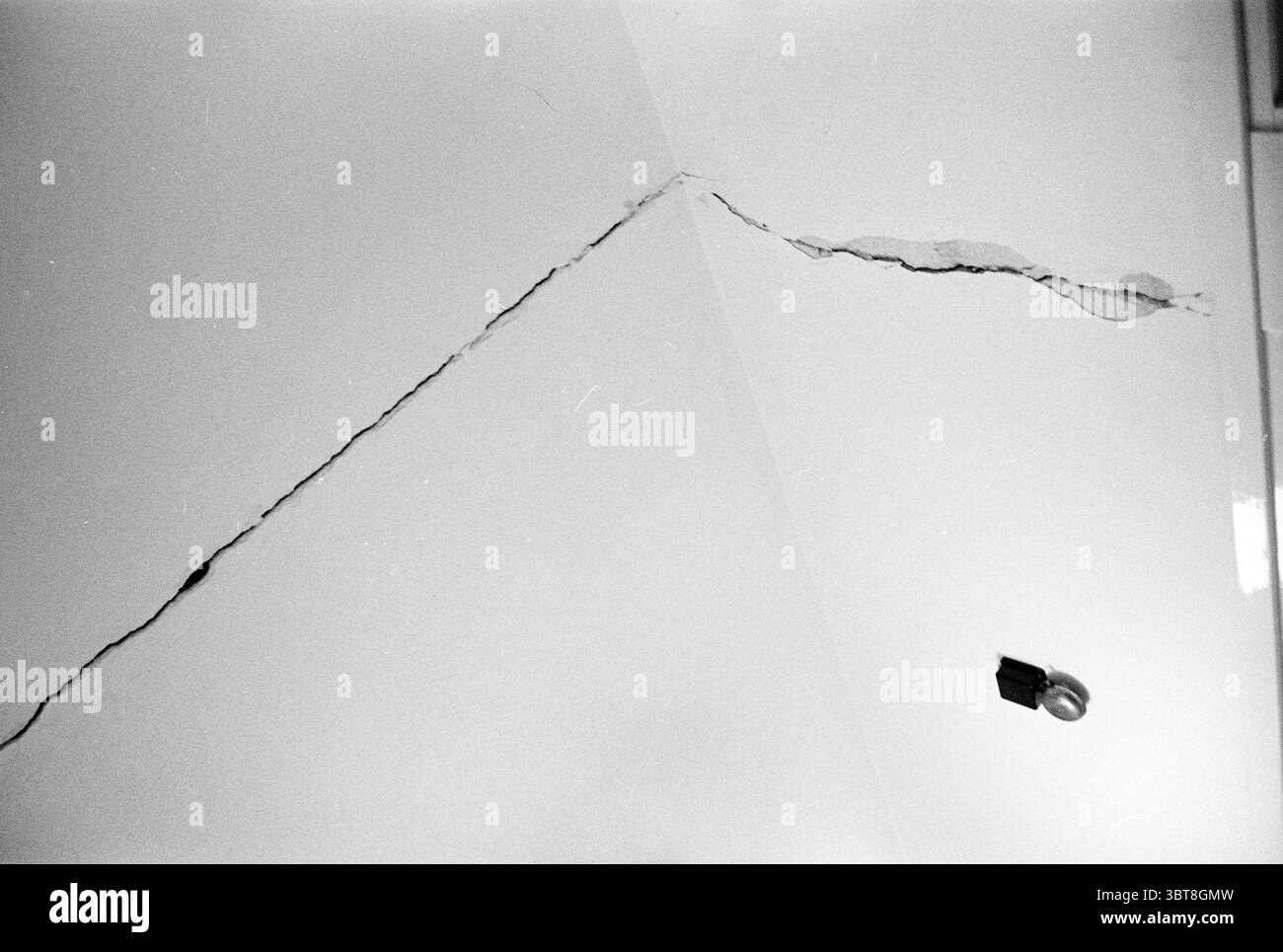 Risse in den Häusern Häuser und Hausbau, Whizgle News, Dutch Desk, Niederlande, 1950 - 2000 am 11-06-1963. Das Bild enthält diese Themen. Die Szene zeigt eine Decke, die von unten betrachtet wird und von einer einzigen Lampe auf der rechten Seite beleuchtet wird. Der Hauptfokus ist ein markanter Riss, der diagonal über die Oberfläche verläuft. Seine zerklüfteten Kanten bilden einen starken Kontrast zur ansonsten glatten Struktur der Decke. Diese Spalte deutet auf Alter und mögliche Vernachlässigung hin, was ein Gefühl der Müdigkeit hervorruft. Die Farbpalette ist gedämpft, dominiert von Grautönen, die zu einem gedämpften Atmo beitragen Stockfoto