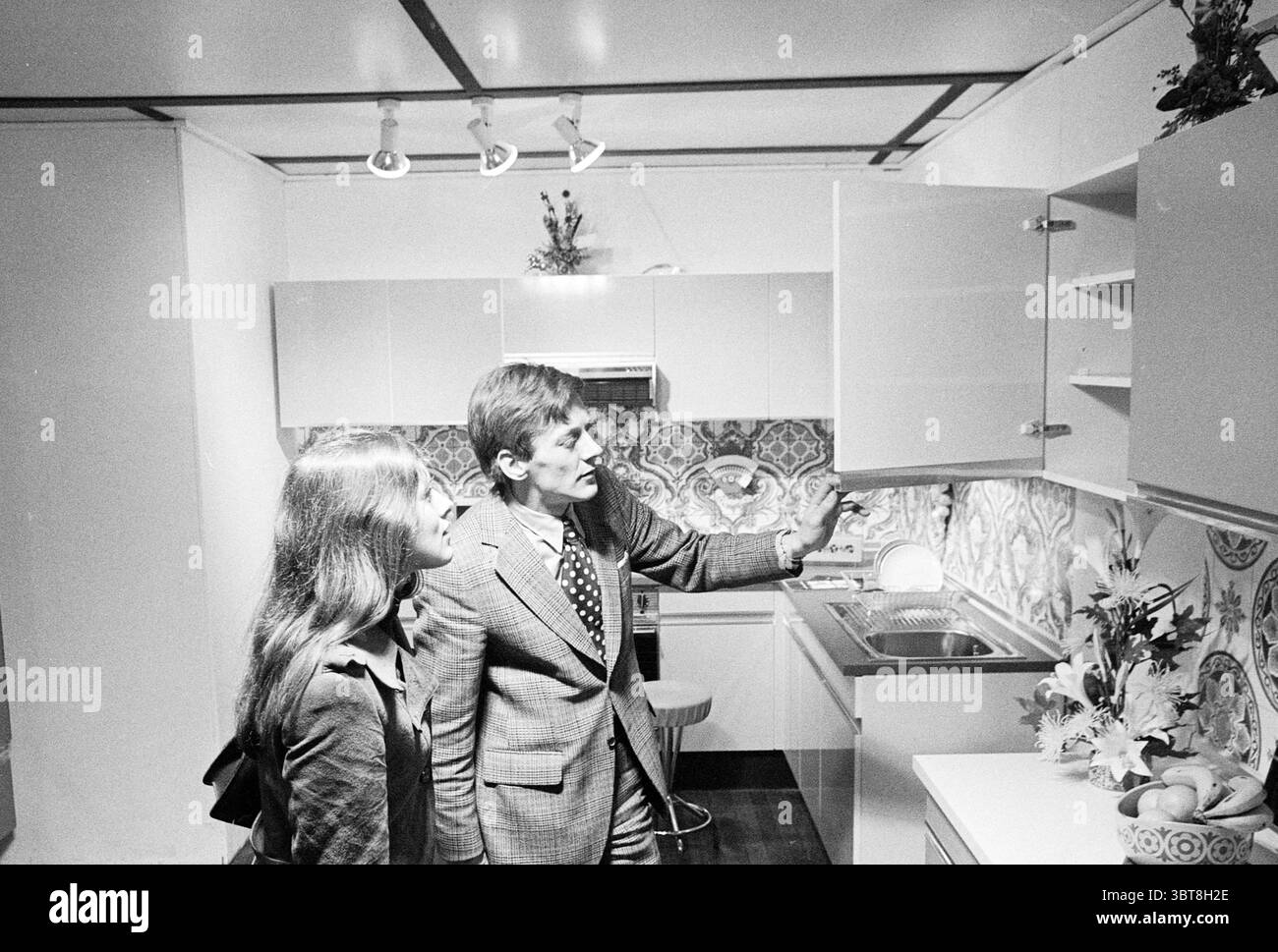 Messe für Herde und Küchen., Whizgle News, Dutch Desk, Niederlande, 1950 - 2000. Dies sind die Elemente im Bild. In einer modernen Kücheneinrichtung führen zwei Personen Gespräche. Der Mann, gekleidet in einem gemusterten Anzug, bewegt sich auf ein offenes Kabinett, sein Ausdruck ist animiert, als er etwas erklärt. Seine Hände, eine hoch und zeigt, deuten darauf hin, dass er ein Detail des Küchendesigns hervorhebt. Die Frau neben ihm, mit langen, glatten Haaren und einem dunklen Outfit, scheint interessiert zu sein und lehnt sich leicht auf ihn zu, während sie die Informationen einnimmt. Die Küche selbst ist es Stockfoto