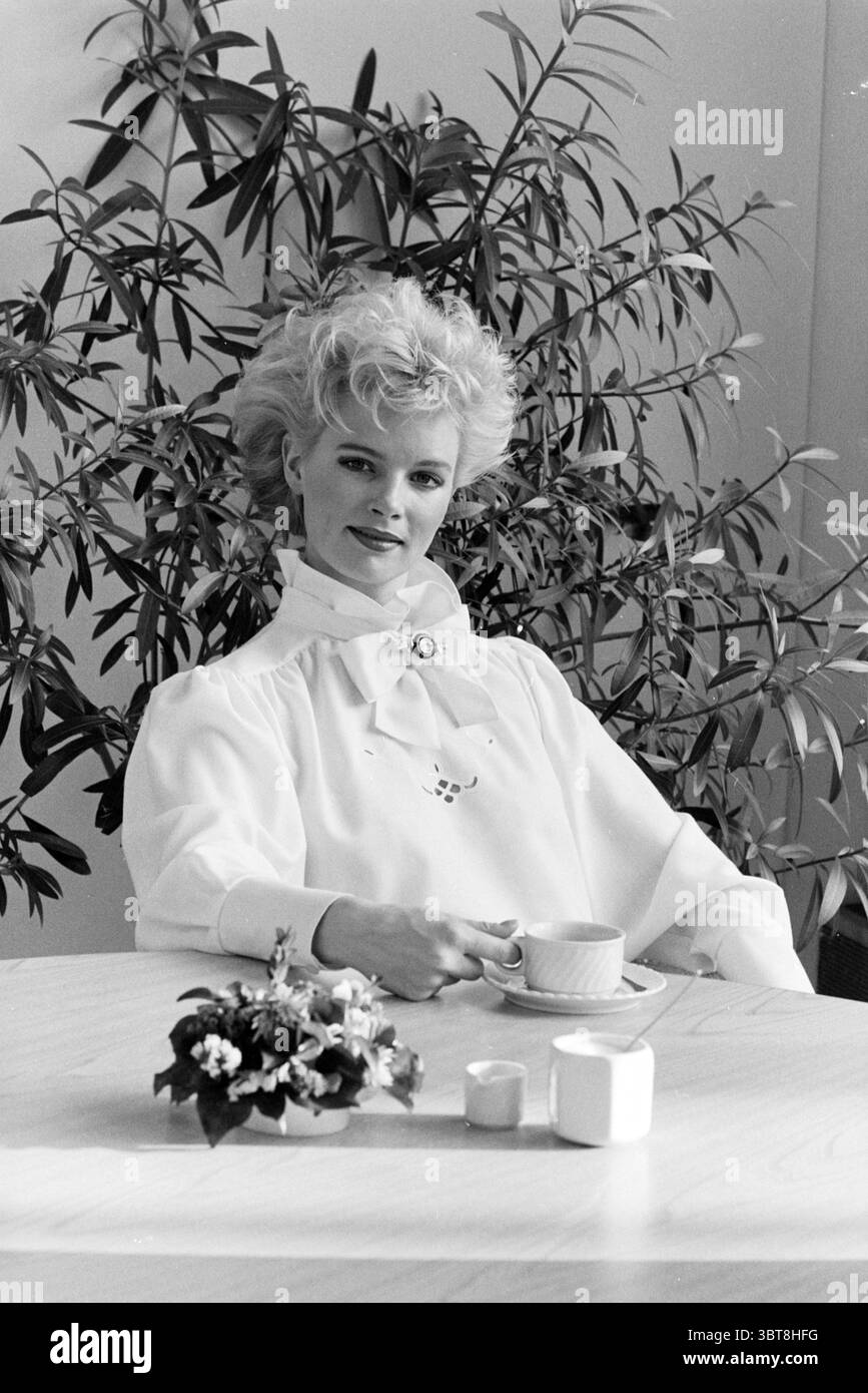Fashion Report Christmas Fashion, Whizgle News, Dutch Desk, Niederlande, 1950 - 2000 am 06-12-1985. Dies sind die Themen im Bild. In einem sanft beleuchteten Innenbereich sitzt eine Frau anmutig an einem runden Holztisch. Ihre Frisur ist voluminös und elegant gestylt, was ihrem Aussehen einen Hauch von Raffinesse verleiht. Sie trägt eine fließende weiße Bluse mit hohem Kragen, die einen schönen Kontrast zu ihren hellen Details bildet. Um sie herum ist eine üppige grüne Pflanze mit langen, schlanken Blättern, die eine ruhige und einladende Atmosphäre schafft. Die leuchtenden Farben der Pflanze unterstreichen die Einfachheit der Szene. Ein Stockfoto