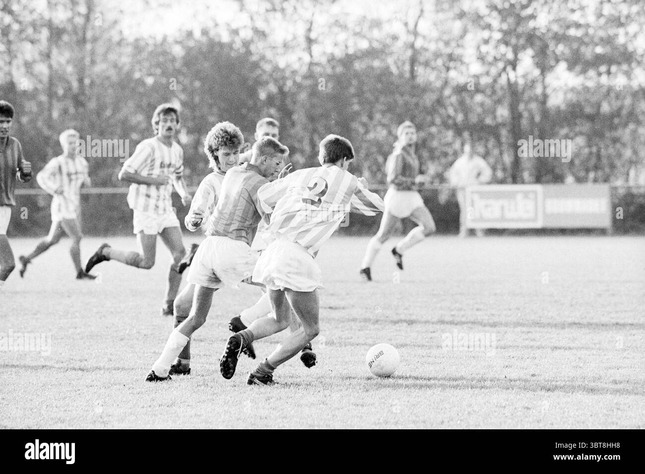 Fußballspiel VV Hillegom - ?, Whizgle News, Dutch Desk, Niederlande, 1950 - 2000 am 05-11-1989. Das Bild enthält diese Themen. In der Szene spielt sich ein dynamisches Fußballspiel auf einem grasbewachsenen Feld ab. Spieler in gestreiften und weißen Trikots sind in einem intensiven Wettkampf mit einem sichtbaren Gefühl der Dringlichkeit in ihren Haltungen. Eine zentrale Figur in einem gestreiften Trikot versucht, einem Verteidiger zu entgehen, der eine solide Nummer auf dem Rücken trägt. Die Farben sind gedämpft; die Trikots zeigen sanfte weiß- und Grautöne, die sich mit dem natürlichen Grün des Feldes und den sanften Brauntönen der Umgebung verbinden Stockfoto