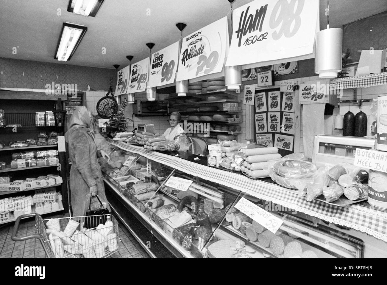 Innenausstattung eines Supermarktes. Whizgle News, Dutch Desk, Niederlande, 1950 - 2000. Dies sind die Themen im Bild. Die Szene zeigt ein traditionelles Feinkost- oder Lebensmittelgeschäft mit Vintage-Schwarz-weiß-Ästhetik. Eine Frau steht an der Theke, in einem langen Mantel gekleidet, mit teilweise bedecktem Kopf, was auf eine kalte Atmosphäre hindeutet. Sie scheint in einem Gespräch mit dem Mitarbeiter hinter der Theke verwickelt zu sein, der eine Schürze trägt und ein einladendes Verhalten zeigt. Die Theke zeigt eine Auswahl an Fleischgerichten, Käse und verschiedenen verpackten Waren, die alle in einem Glas angeordnet sind Stockfoto