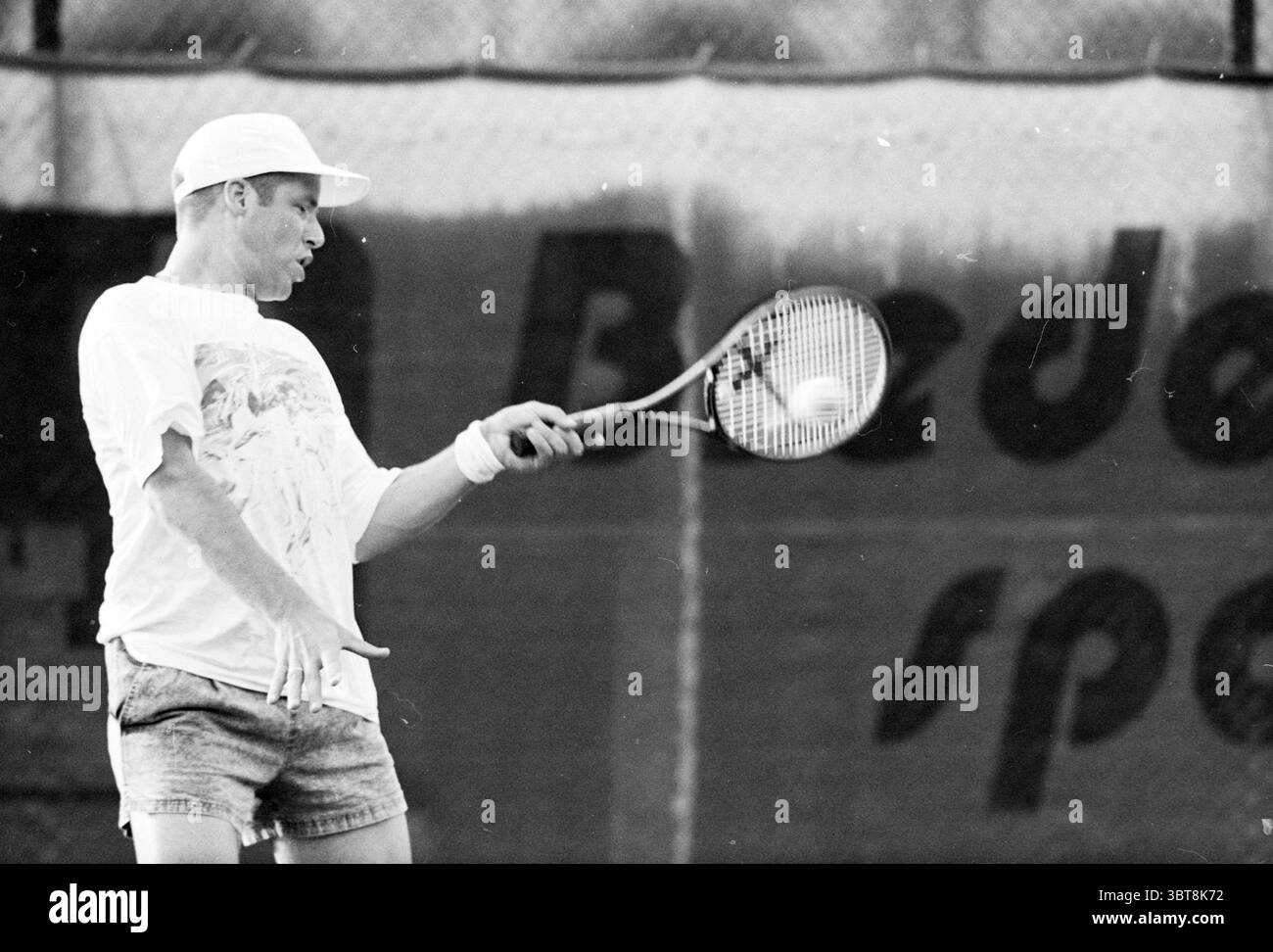 Tennis Brederode v.d. Mountain, Whizgle News, Dutch Desk, Niederlande, 1950 - 2000 am 12-07-1995. Dies sind die Elemente im Bild. In einer dynamischen Sportszene wird ein Tennisspieler mitten im Schwung erfasst, während er sich auf den Ball vorbereitet. Er trägt ein weites weißes T-Shirt mit farbenfrohem grafischem Design, das einen schönen Kontrast zu seinen dunkleren, leicht verblassten Shorts bildet. Die Shorts sind stylisch kurz und sorgen so für eine energiegeladene Atmosphäre. Der Spieler hat einen fokussierten Ausdruck, der auf Konzentration und Entschlossenheit hinweist. Seine Haltung ist athletisch, mit einem Fuß für ihn positioniert Stockfoto