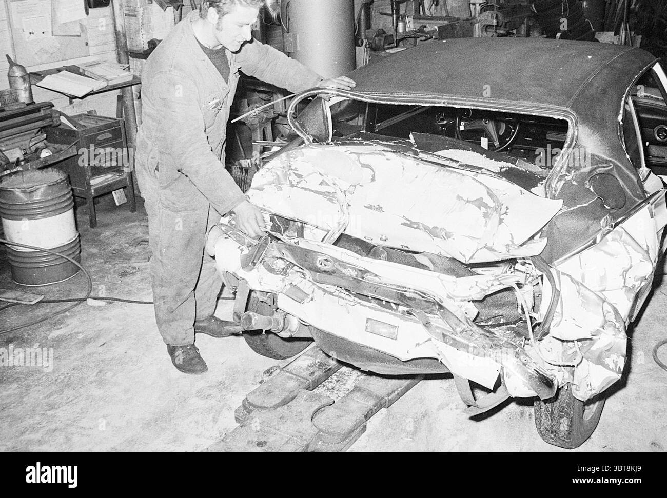 Mann in der Nähe eines Autounfalls in der Garage. Whizgle News, Dutch Desk, Niederlande, 1950 - 2000. Diese Themen sind in der Abbildung dargestellt. In einer schwach beleuchteten Werkstatt, die von einer rauchigen Atmosphäre erfüllt ist, wird ein Mann in einem leicht getragenen grauen Jumpsuit gesehen, der intensiv an einem beschädigten Fahrzeug arbeitet. Die Szene wird vom zerknitterten Heck eines Autos dominiert, das in der Mitte deutlich positioniert ist. Die Karosserie des Fahrzeugs besteht aus kräftigem weiß und mattem Grau, mit deutlichen Falten und Dellen, die auf eine starke Stoßbelastung hinweisen. Der Mann mit kurzen, abgeschnittenen Haaren zeigt einen entschlossenen Ausdruck, während er sich nach vorne neigt, um den Schaden zu beurteilen Stockfoto