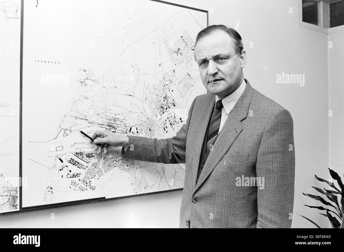 Mr. Van Tol Etching Fields Hoogovens Hoogovens, Whizgle News, Dutch Desk, Niederlande, 1950 - 2000 am 23-01-1985. Diese Themen sind in der Abbildung dargestellt. In der Szene steht ein Mann selbstbewusst in einer Büroumgebung und bewegt sich auf eine große Karte, die an der Wand hinter ihm montiert ist. Er trägt einen klassischen, maßgeschneiderten Anzug mit Krawatte, der einen Hauch von Formalität und Professionalität vermittelt. Die Karte selbst ist detailliert und weist verschiedene Linien und Markierungen auf, die darauf hindeuten, dass sie sich auf Stadtplanung oder Geographie bezieht, wobei eine Mischung aus hellen und dunklen Schattierungen verschiedene Bereiche anzeigt. Der Backgr Stockfoto