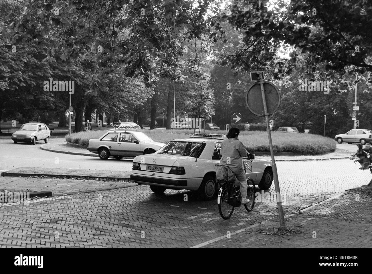 Kenaupark Roundabout Haarlem Kenaupark the Netherlands, Whizgle News, Dutch Desk, Niederlande, 1950 - 2000 am 13-09-1990. Diese Themen sind in der Abbildung dargestellt. In dieser Szene wird ein kreisförmiger Verkehrsbereich von einem üppigen Baumdach eingerahmt, deren Blätter ein abwechslungsreiches Licht- und Schattenmuster auf dem Boden erzeugen. Die gesamte Farbpalette ist monochromatisch und hebt den Kontrast von tiefen Schwarztönen und sanften Grautönen in der gesamten Einstellung hervor. Vor der Szene steht ein Radfahrer in heller Kleidung, der die kreisförmige Kreuzung durchquert. Das Fahrrad ist einfach und hat ein klassisches Design Stockfoto