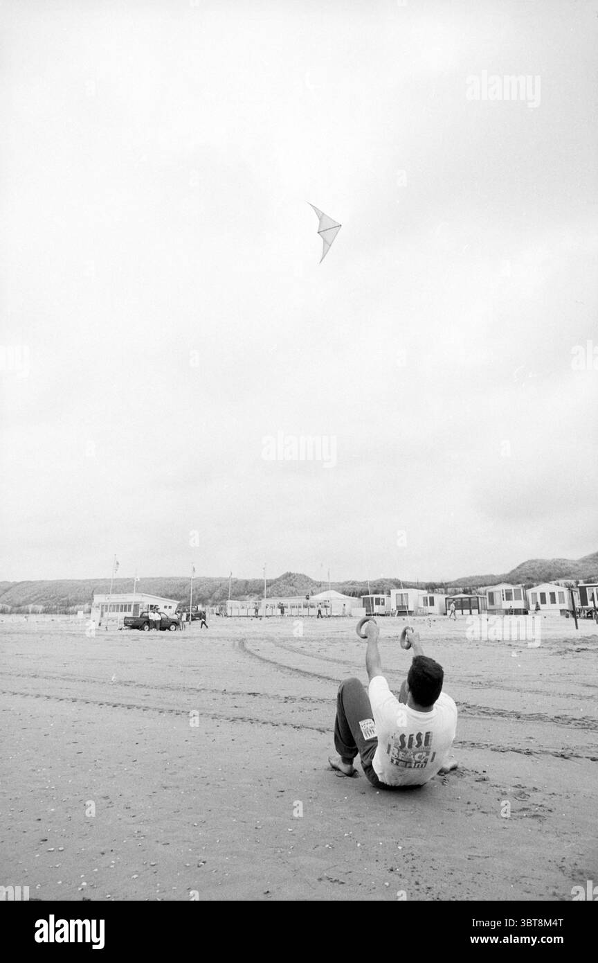 Drachenfliegen Demo Wijk aan Zee Wijk aan Zee, Whizgle News, Dutch Desk, Niederlande, 1950 - 2000 am 17.07.1992. Dies sind die Themen im Bild. Die Szene entfaltet sich an einem Sandstrand unter bewölktem Himmel und schafft eine gedämpfte Atmosphäre. Ein Mann sitzt auf dem Sand und lehnt sich zurück mit seinem Gewicht an einer Hand. Er trägt ein helles Hemd, im Kontrast zu den dunkleren Sandtönen. Sein fokussierter Ausdruck zeigt seine Freude, wenn er anmutig einen farbenfrohen Drachen in die Luft wirft. Der Drachen selbst ist ein markantes Element, das mit einer lebendigen Farbpalette darüber gleitet, die einen Hauch von Farbe erzeugt Stockfoto