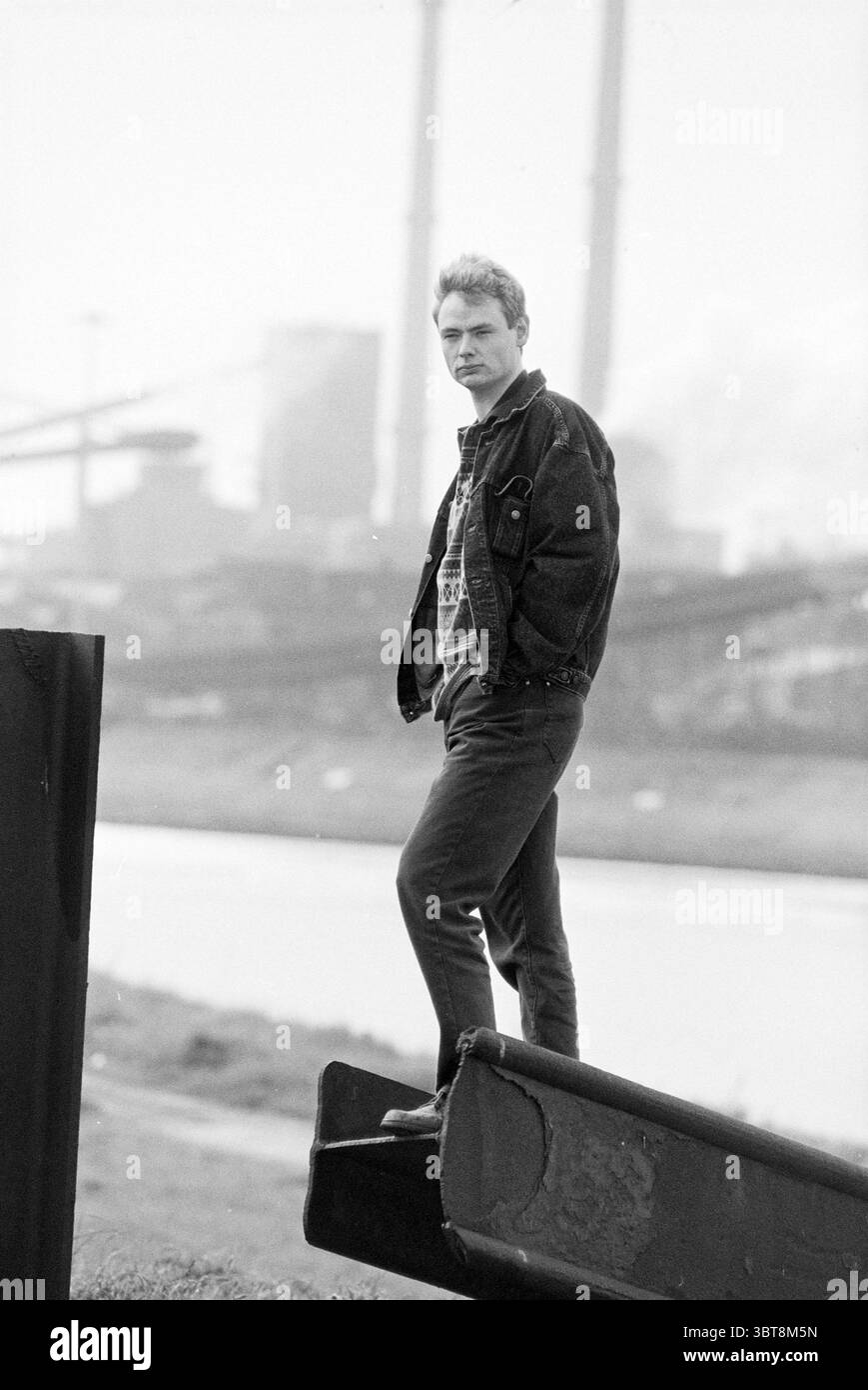 Herr Bal CNV Velsen-Noord Velsen-Noord, Whizgle News, Dutch Desk, Niederlande, 1950 - 2000 am 16-05-1994. Diese Themen sind in der Abbildung dargestellt. Die Szene zeigt einen jungen Mann, der selbstbewusst auf einem Stück Industrieschutt steht, vielleicht einem verrosteten Balken oder Rohr, der sich am Rand eines Wasserkörpers befindet. Er trägt eine Denim-Jacke über einem grafischen T-Shirt und eine taillierte Hose, die für einen lässigen, aber stilvollen Look sorgt. Sein Haarschnitt ist kurz und leicht zerzaust, was sein jugendliches Verhalten noch verstärkt. Im Hintergrund ist eine von Industriestrukturen dominierte Landschaft sichtbar, einschließlich Stockfoto