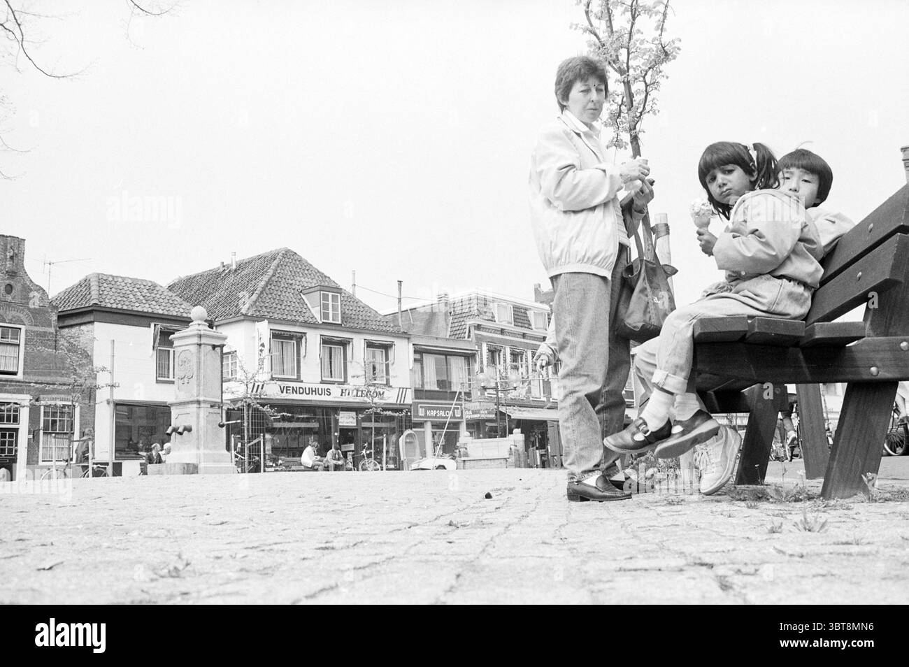 Zentrum Hillegom Menschen auf Bänken in der Sonne Hillegom Sommer Bilder Hillegom, Whizgle News, Dutch Desk, Niederlande, 1950 - 2000 am 08-05-1985. Diese Themen werden in der Abbildung angezeigt. Auf einem belebten Stadtplatz steht eine Frau im Vordergrund, die ein kleines Bündel hüllt, während sie zur Seite blickt. Sie trägt eine helle Jacke und hat kurze, dunkle Haare, die eine Aura der Aufmerksamkeit ausstrahlen. Neben ihr sitzen zwei Kinder auf einer Holzbank und teilen sich einen süßen Leckerbissen, wobei ein Kind auf die Frau blickt, während das andere sich auf ihren Snack konzentriert. Der Hintergrund zeigt einen charmanten Stockfoto