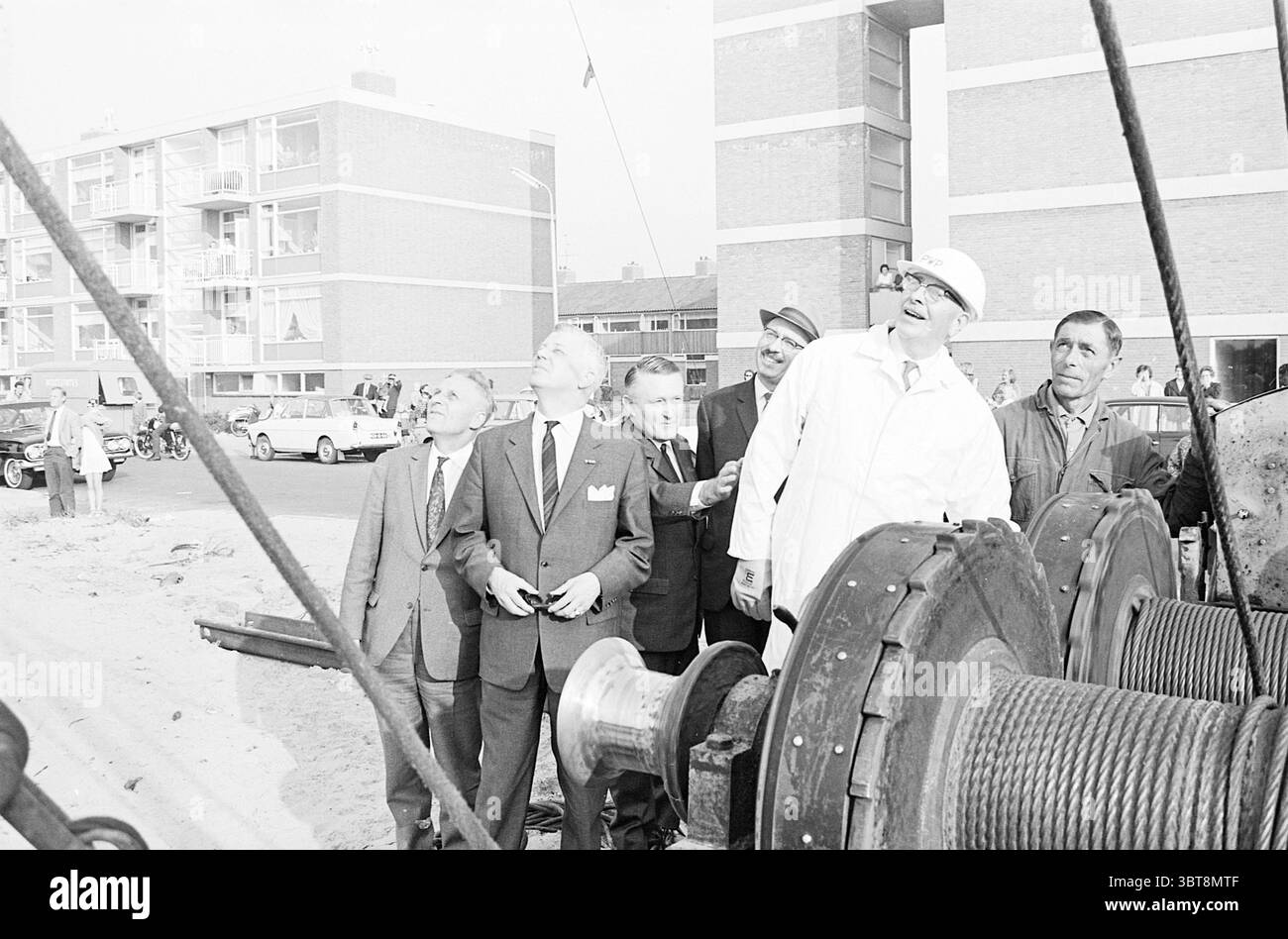 Die erste Staffel für ein Projekt., Whizgle News, Dutch Desk, Niederlande, 1950 - 2000. Dies sind die Elemente im Bild. In einer geschäftigen Umgebung im Freien steht eine Gruppe von Männern aufmerksam, ihre Blicke nach oben gerichtet. Sie sind in formeller Kleidung gekleidet, die meisten in Anzügen und Krawatten, was ein Gefühl von Anlass oder Wichtigkeit widerspiegelt. Eine Person zeichnet sich in einem weißen Mantel und einem Schutzhelm aus und strahlt eine Atmosphäre von Autorität oder Fachwissen aus. Im Hintergrund befindet sich ein flaches Gebäude mit mehreren rechteckigen Fenstern, die auf eine städtische Umgebung hinweisen. Autos parken entlang der Straße, was auf ein geschäftiges Treiben hindeutet Stockfoto