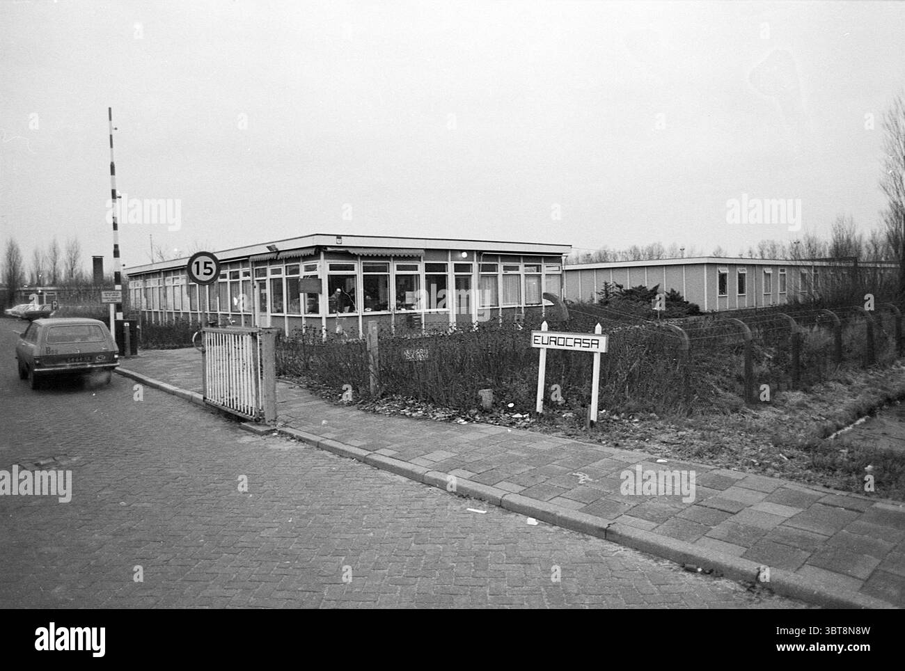 Eurocasa Eingangstor, Whizgle News, Dutch Desk, Niederlande, 1950 - 2000 am 12-1976. Das Bild enthält diese Themen. Die Szene präsentiert eine ruhige, urbane Umgebung, die eine etwas öde Gegend zu sein scheint. Ein niedriges, einstöckiges Gebäude mit modernem architektonischen Stil ist hervorzuheben, das durch große Fenster gekennzeichnet ist, die einen Blick auf das Innere erlauben. Die Fassade ist größtenteils horizontal, mit einem flachen Dach und einer ordentlichen Anordnung von Glaspaneelen. Die Farbpalette ist gedämpft und überwiegend grau, was auf eine düstere Umgebung hindeutet. Im Vordergrund verläuft parallel dazu eine gut gepflegte Straße Stockfoto