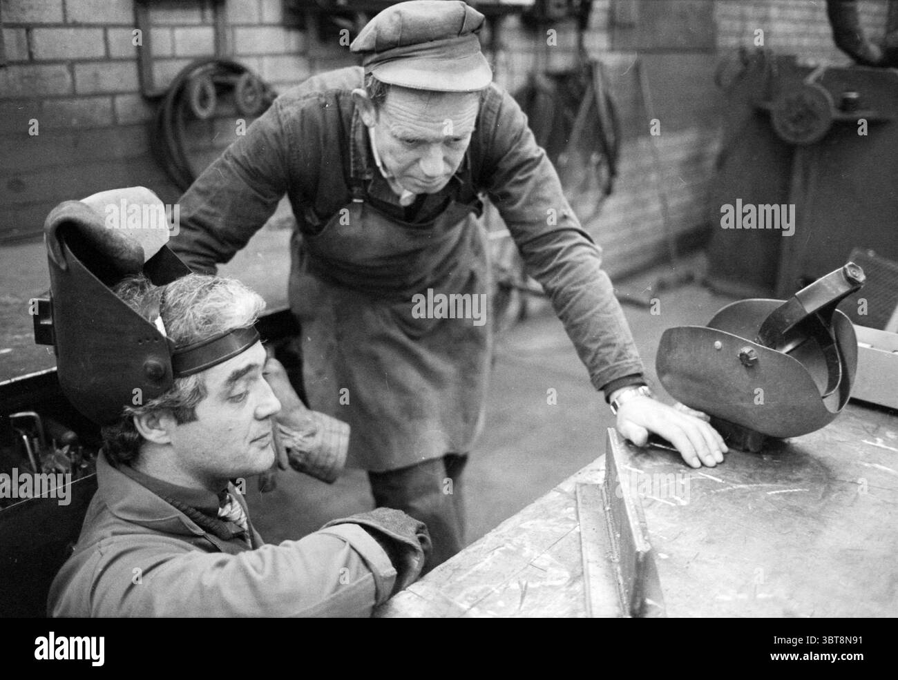 AGA-Gas melden. Support Group B.V., Whizgle News, Dutch Desk, Niederlande, 1950 - 2000 am 09-02-1983. Dies sind die Elemente im Bild. In einer schwach beleuchteten Werkstatt führen zwei Männer über eine Werkbank ein fokussiertes Gespräch. Die Umgebung zeichnet sich durch ihre industrielle Umgebung aus, die mit rauen Texturen und zweckmäßigen Werkzeugen verstreut ist. Der erste sitzende Mann trägt eine dunkle Kappe und einen Kopfschmuck, der für Schweißen oder Metallarbeiten typisch ist. Sein Ausdruck ist Konzentration, was darauf hindeutet, dass er sich entweder mitten in einer Aufgabe befindet oder Informationen aufnimmt. Seine Kleidung ist praktisch für die Arbeit Stockfoto