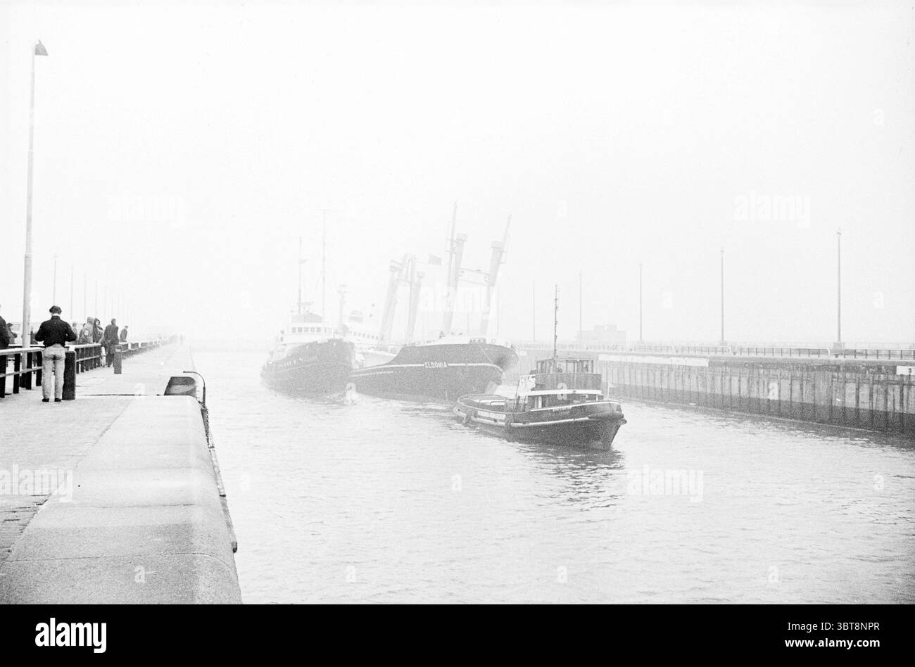 Französische Frachter Eldonia schwer beschädigt in Noordersluis-Schiffen, Whizgle News, Dutch Desk, Niederlande, 1950 - 2000 am 29.12.1963. Das Bild enthält diese Themen. In dieser Szene wird die Umgebung durch den dicken Nebel, der die Sicht verdeckt, von einem geheimnisvollen Gefühl umhüllt. Die gesamte Farbpalette ist gedämpft, dominiert von Grau- und Schwarztönen, was eine düstere, fast nostalgische Atmosphäre schafft. Auf einer Seite ist ein Industriegebiet mit einem stabilen Gehweg gesäumt. Einige wenige Figuren stehen lässig am Rand, ihre Silhouetten sind durch den Dunst leicht verwischt. Sie scheinen hinauszuschauen Stockfoto