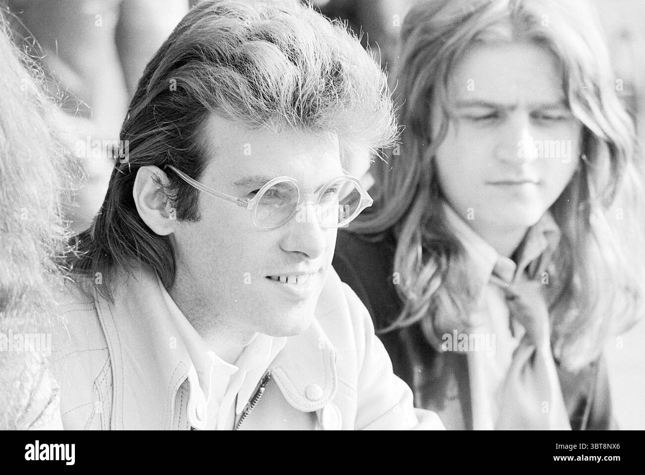 Portraits Roxy Music Portraits, Whizgle News, Dutch Desk, Niederlande, 1950 - 2000 am 27-05-1973. Die Abbildung zeigt diese Themen. In der Szene sitzen zwei Personen dicht beieinander, die beide mit ihren unterschiedlichen Frisuren und Ausdrucksformen Aufmerksamkeit erregen. Der linke trägt einen voluminösen, gestylten Haarschnitt mit markanten, fließenden Locken, die sein Gesicht umrahmen. Seine Brille ist rund und übergroß und verleiht einen Hauch von Intellektualität oder künstlerischem Flair. Er trägt eine helle Jacke, die einen subtilen Kontrast zu den sanfteren Haaren und Teint bildet. Sein Ausdruck ist Contemplati Stockfoto