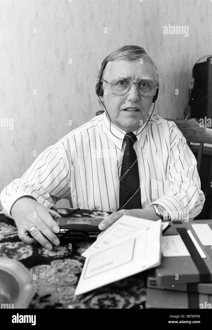 Mr Drijver Sound Zeitung IJmuiden Sound People Zeitungen und Zeitschriften IJmuiden the Netherlands, Whizgle News, Dutch Desk, Niederlande, 1950 - 2000 am 23-02-1990. Diese Themen sind in der Abbildung dargestellt. Die Szene zeigt einen älteren Mann, der an einem Tisch sitzt und ein Gefühl von Autorität und Konzentration ausstrahlt. Er trägt ein knuspriges, weißes Hemd mit dünnen, vertikalen blauen Streifen, ergänzt durch eine dunkle Krawatte mit dezentem Muster, die sein professionelles Aussehen unterstreicht. Seine Brille mit einem etwas dicken Rahmen liegt bequem auf seiner Nase und verleiht ihm einen fleißigen Blick. Er ist mit Kopfhörern ausgestattet, Stockfoto