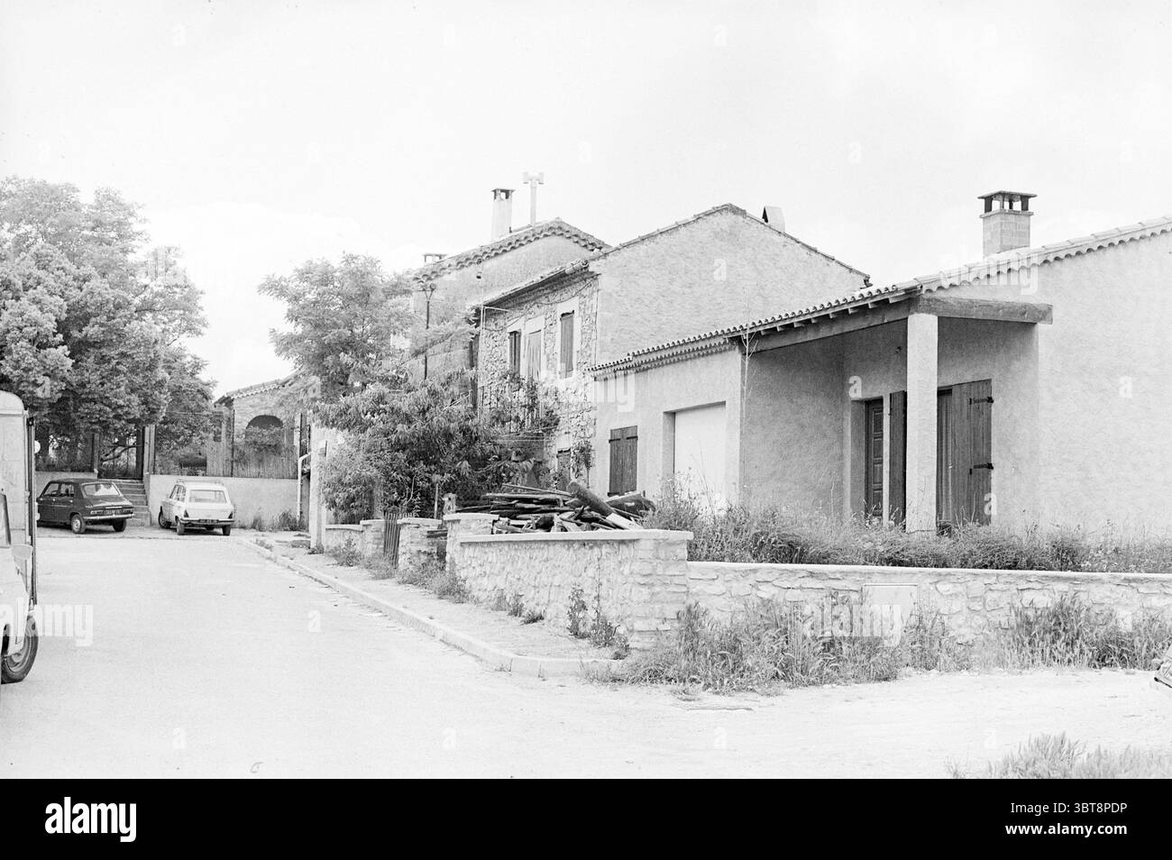 Bericht Mesjaunne France - Solar Panels for Heat Report, Whizgle News, Dutch Desk, Niederlande, 1950 - 2000 am 17-06-1975. Diese Themen sind in der Abbildung dargestellt. Die Szene zeigt eine ruhige Straße, die von Wohnhäusern gesäumt ist. Die Architektur spiegelt einen rustikalen Charme wider, mit Häusern aus Stein und hellem Stuck. Einige Gebäude sind mit Holzläden ausgestattet, die teilweise geschlossen sind und eine einladende und dennoch ruhige Atmosphäre vermitteln. Auf der linken Seite parken einige Fahrzeuge entlang der Straße – ein Van und ein kleineres Auto, beide in gedämpften Tönen, die sich harmonisch mit der Umgebung harmonieren. Die Fahrzeuge Stockfoto