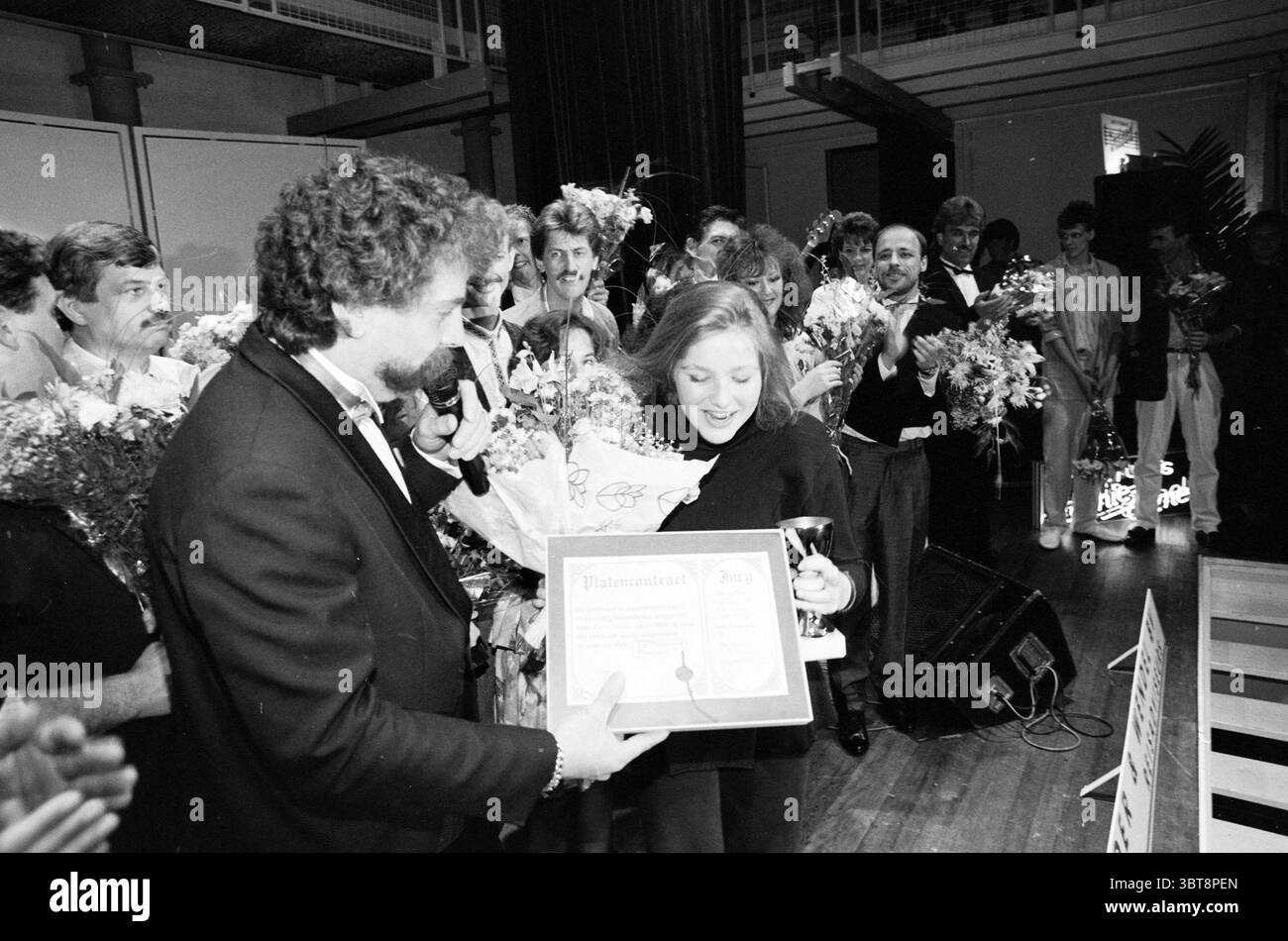 Haarlem Gesangsfestival Finale Festival Festivitäten und Manifestationen Gesangschöre und Gesangschöre Haarlem the Netherlands, Whizgle News, Dutch Desk, Niederlande, 1950 - 2000 am 12-05-1989. Dies sind die Elemente im Bild. Die Szene ist voller Aufregung und Feiern. Im Vordergrund steht ein Mann in einem Smoking, der ein Mikrofon hält und sich mit der versammelten Menge beschäftigt. Sein Haar ist lockig und voluminös, was seinem Aussehen ein dynamisches Element verleiht. Neben ihm strahlt eine Frau vor Freude, während sie ein gerahmtes Zertifikat besitzt, wahrscheinlich eine bedeutende Auszeichnung oder Anerkennung. Um diese t Stockfoto