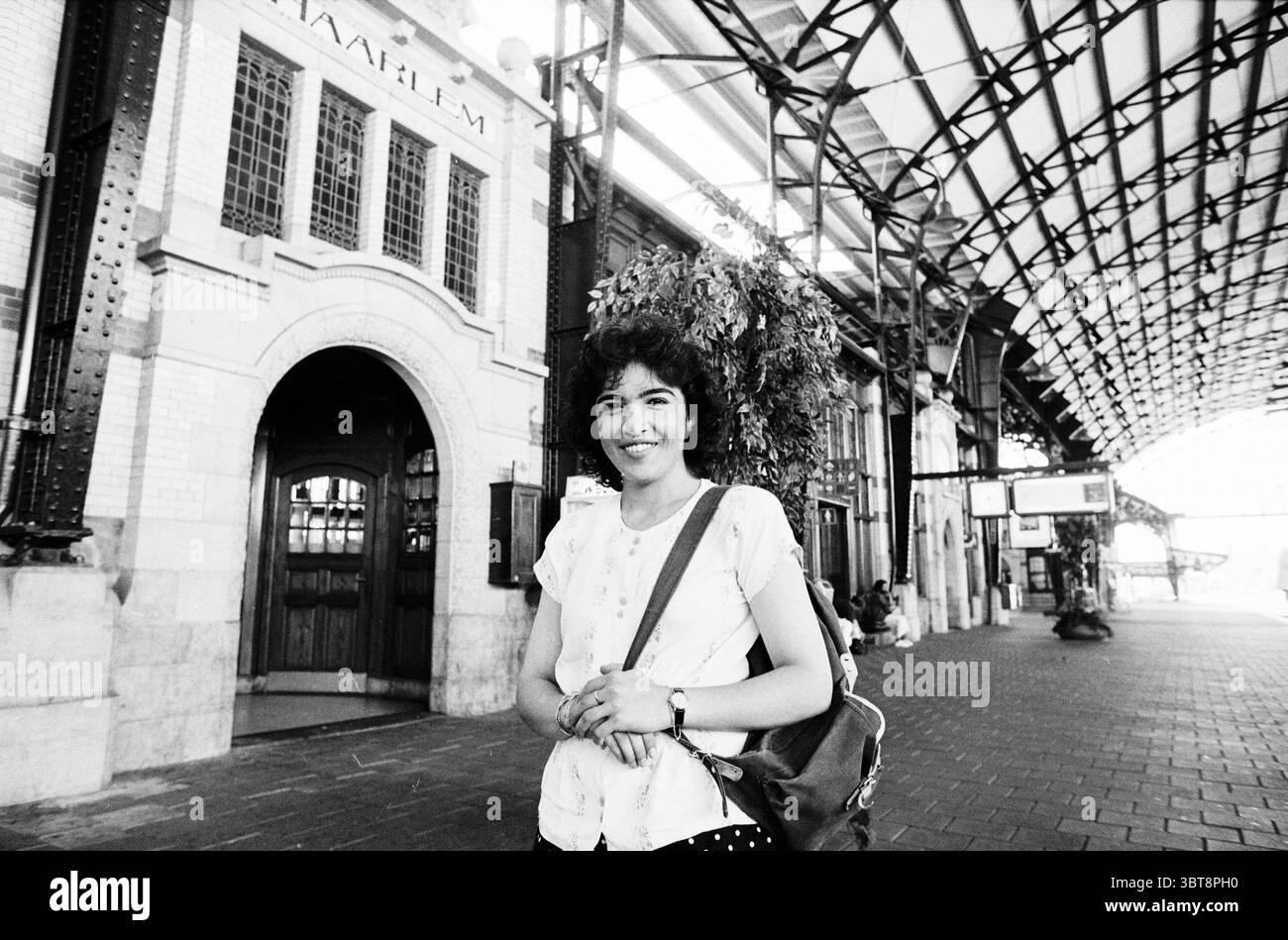 Dame auf dem Bahnsteig am Bahnhof Haarlem, Whizgle News, Dutch Desk, Niederlande, 1950 - 2000 am 14-08-1997. Das Bild enthält diese Themen. Eine Frau steht selbstbewusst in einem belebten Bahnhof, der sich durch seine architektonische Eleganz und seine komplizierten Details auszeichnet. Sie hat lockige Haare und ein warmes Lächeln, was eine einladende Aura schafft. Ihr Outfit ist leicht und lässig. Sie verfügt über ein weißes Oberteil mit kleinen Mustern und einen dunklen Rock, der Kontrast setzt. Der Bahnhof hat eine hohe, bogenförmige Decke, die mit Metallbalken gesäumt ist, was ihm eine geräumige Atmosphäre verleiht. Die Backsteinmauern sind mit großen, hohen Fenstern versehen Stockfoto