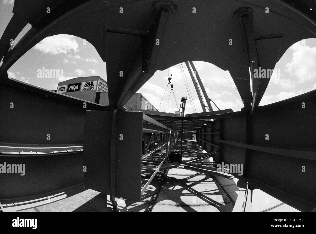 Crane at Figee, Whizgle News, Dutch Desk, Niederlande, 1950 - 2000 am 22-05-1991. Die Abbildung zeigt diese Themen. Die Szene präsentiert eine industrielle Umgebung, die durch Stahlträger in geometrischem Rahmen gekennzeichnet ist. Die Perspektive ist niedrig und abgewinkelt und erfasst das komplizierte Gitter von Metallarbeiten, die sich in Richtung eines zentralen Brennpunkts konvergieren. Die in einem gedämpften Grauton lackierten Balken weisen einen metallischen Glanz auf, der das Umgebungslicht reflektiert und ihr robustes Erscheinungsbild unterstreicht. Oben offenbart sich der Himmel durch die Lücken in der Struktur und zeigt ein helles azurblaues, von Grippe durchzogenes Licht Stockfoto